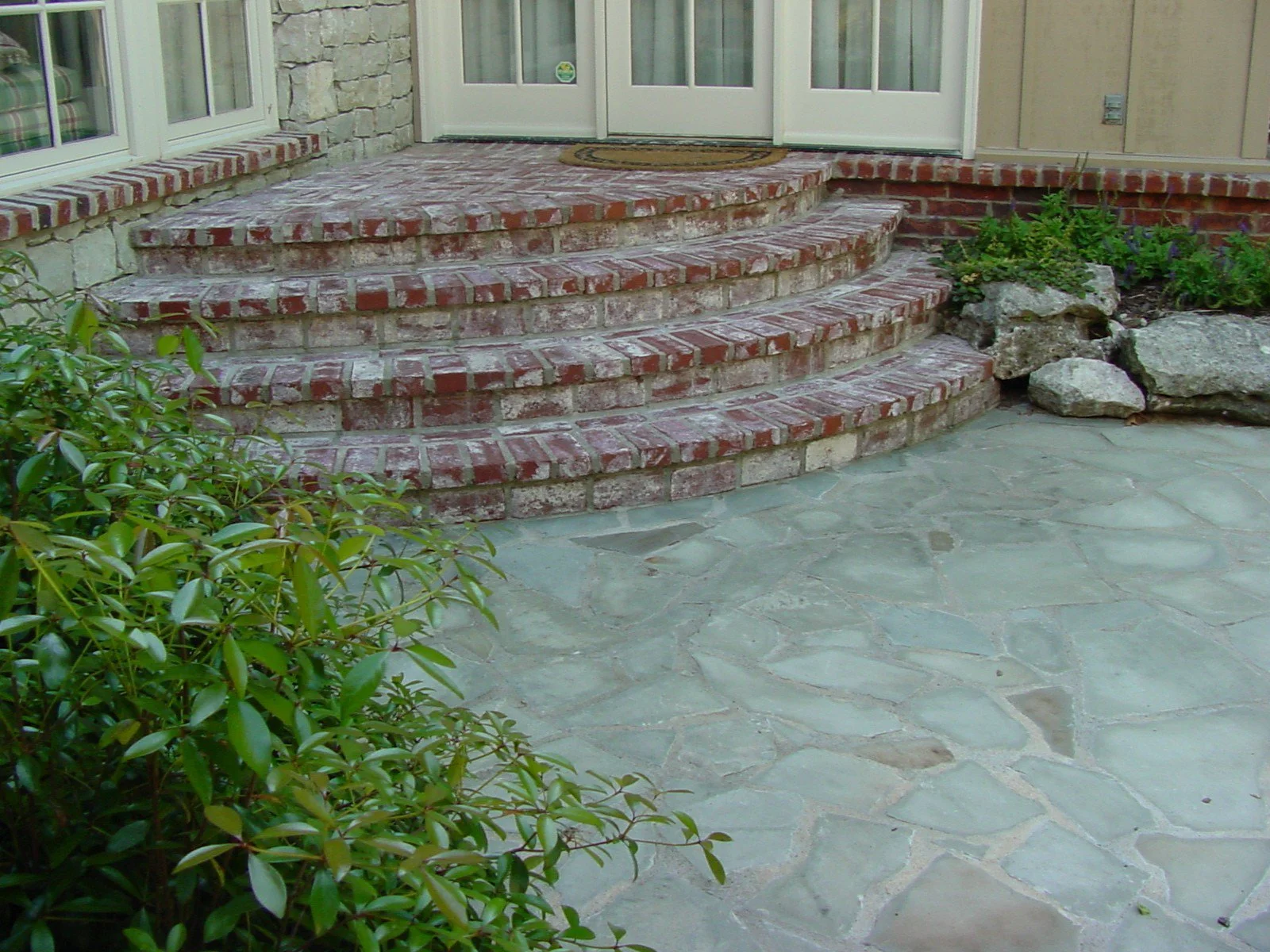 Brick steps leading up to a door with glass panels, featuring green plants and rocks on either side, and a stone patio in the foreground.