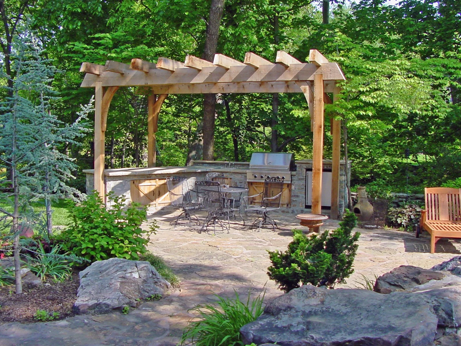 Outdoor patio area with a wooden pergola, metal chairs, a stone barbecue, a wooden bench, and lush green trees and plants surrounding the space.