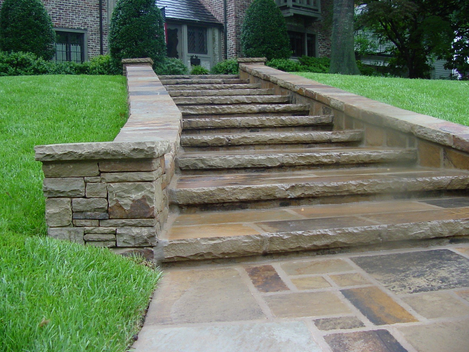 A stone staircase with a stone railing leading up to a brick house. The staircase is surrounded by green grass and bushes, with trees in the background.