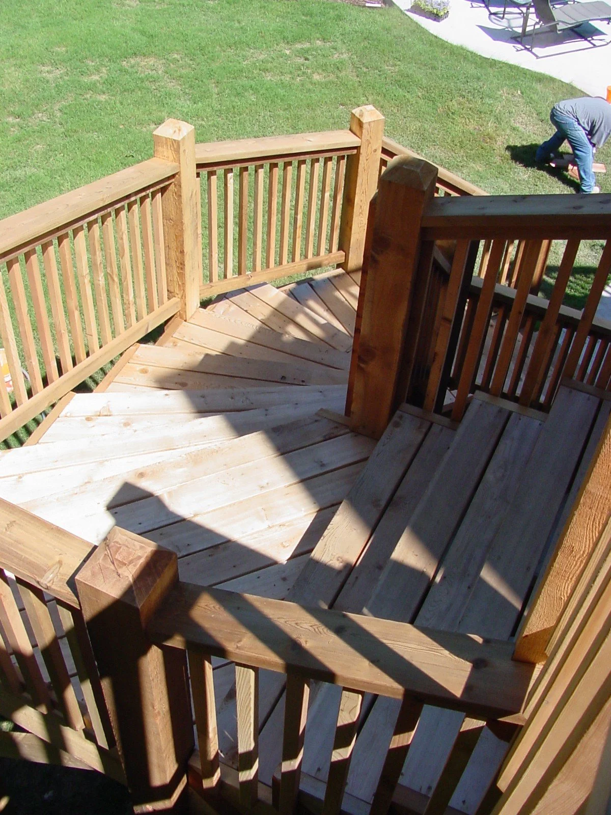 A wooden staircase leading down to a grassy yard with a shadow cast on the steps, and part of an outdoor patio with lounge chairs in the background.