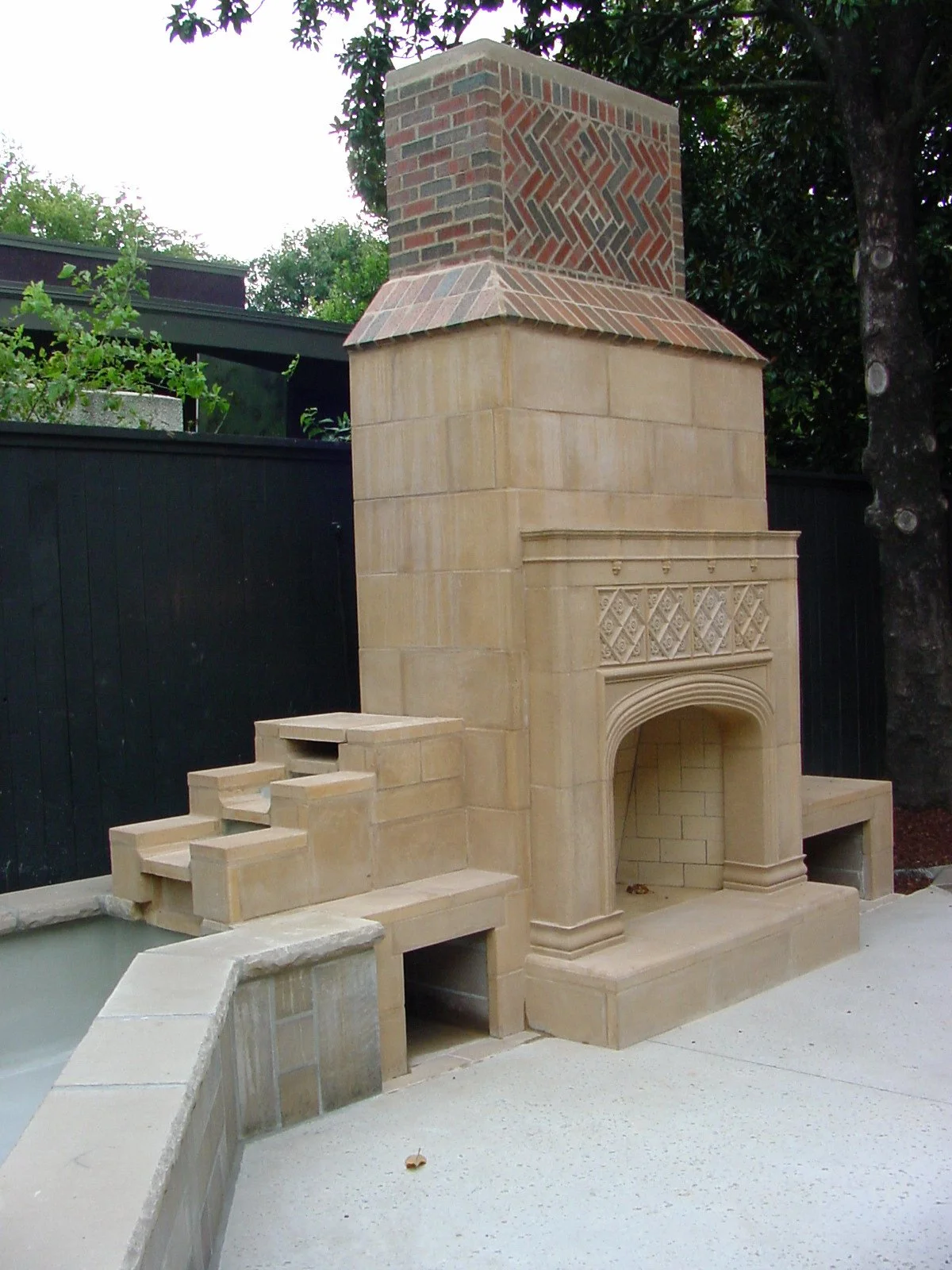 Outdoor stone fireplace with decorative brick chimney, surrounded by a concrete patio, next to a black wooden fence and green trees.