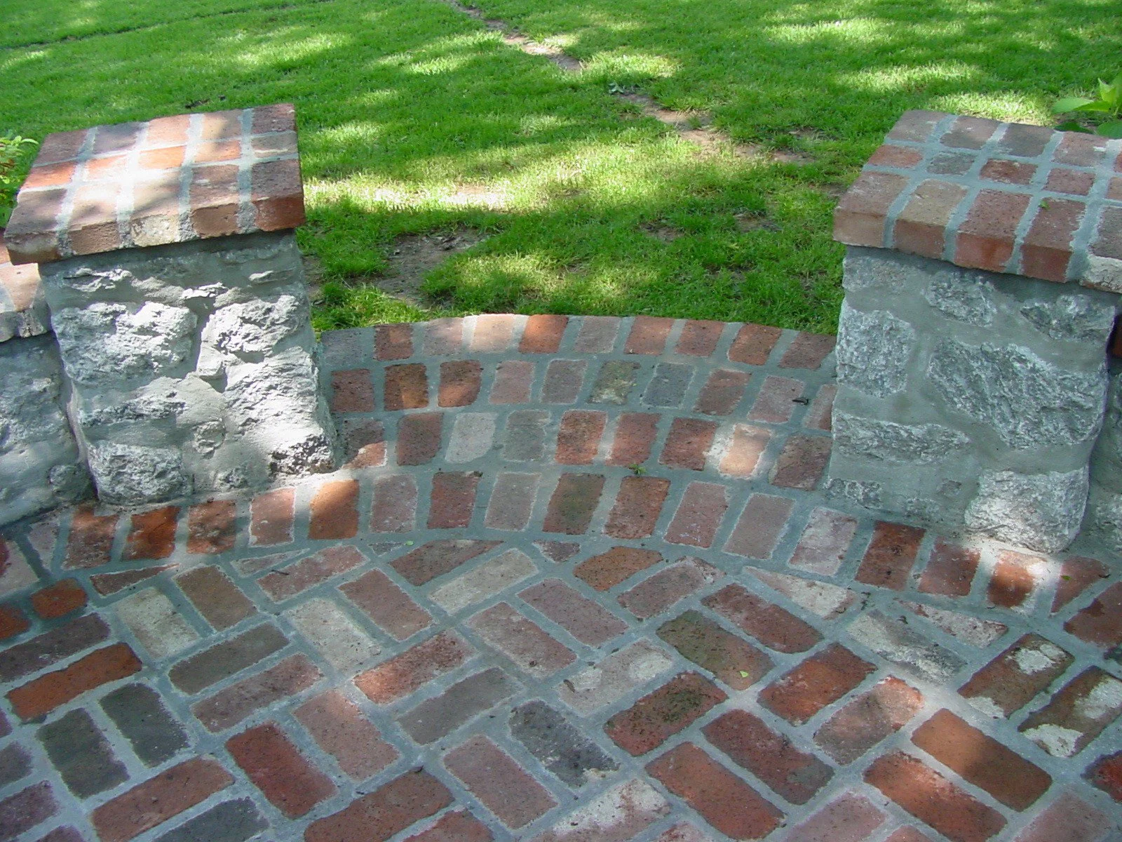 Close-up of a brick and stone decorative bench with brick pillars and curved brick seating area, surrounded by green grass and shaded sunlight.