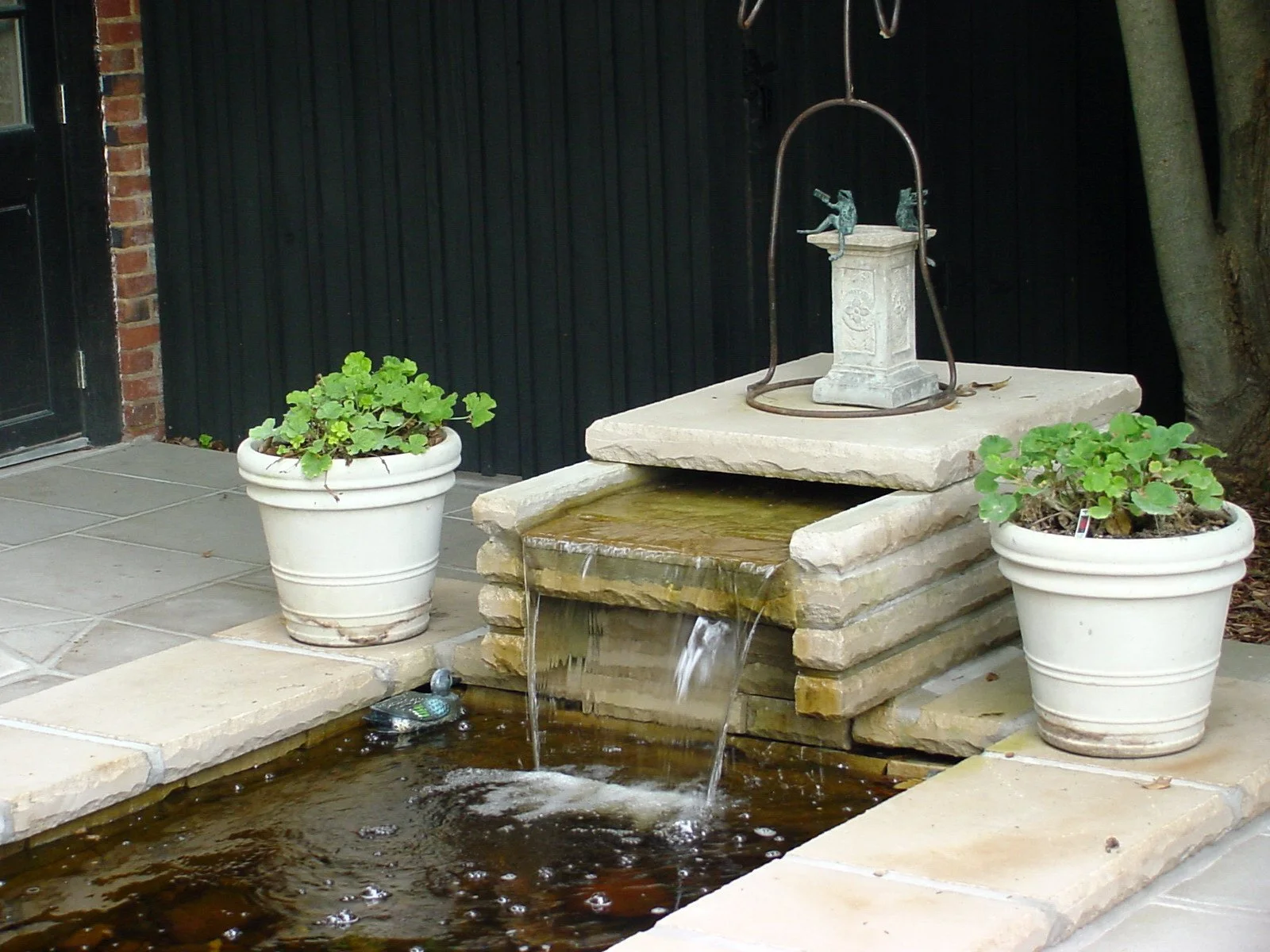 Outdoor decorative water fountain with a tiered stone structure, water flowing into a small pond, and potted plants on each side.
