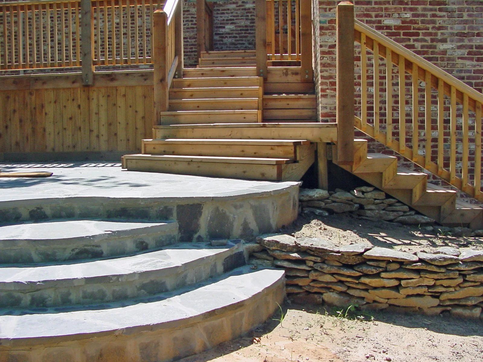 Wooden stairs leading up to a house with brick walls, with a stone-lined patio area and a curved concrete staircase in the foreground.