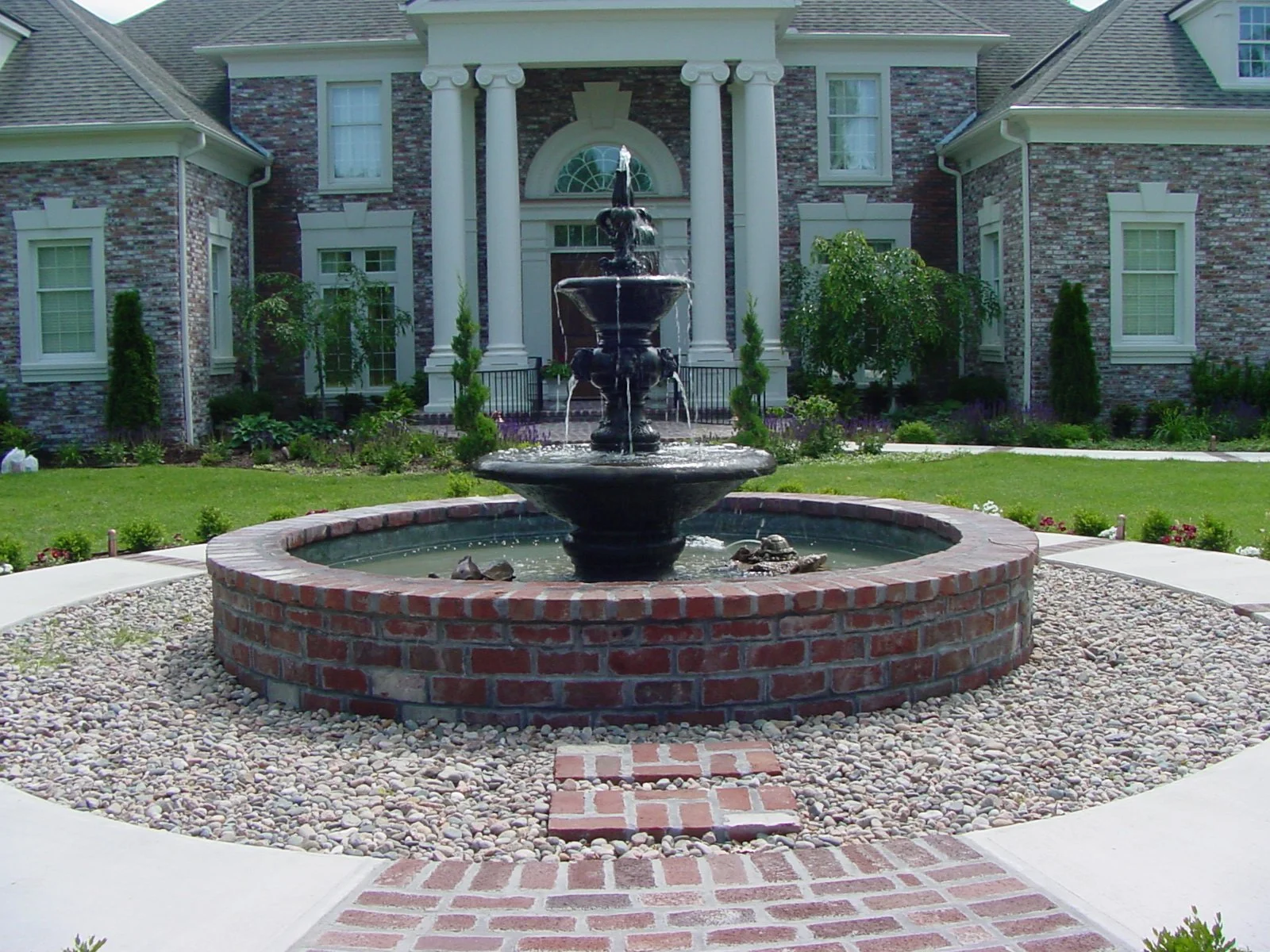 Front yard with a circular brick and stone fountain, green lawn, shrubbery, and a large brick house with columns and multiple windows.