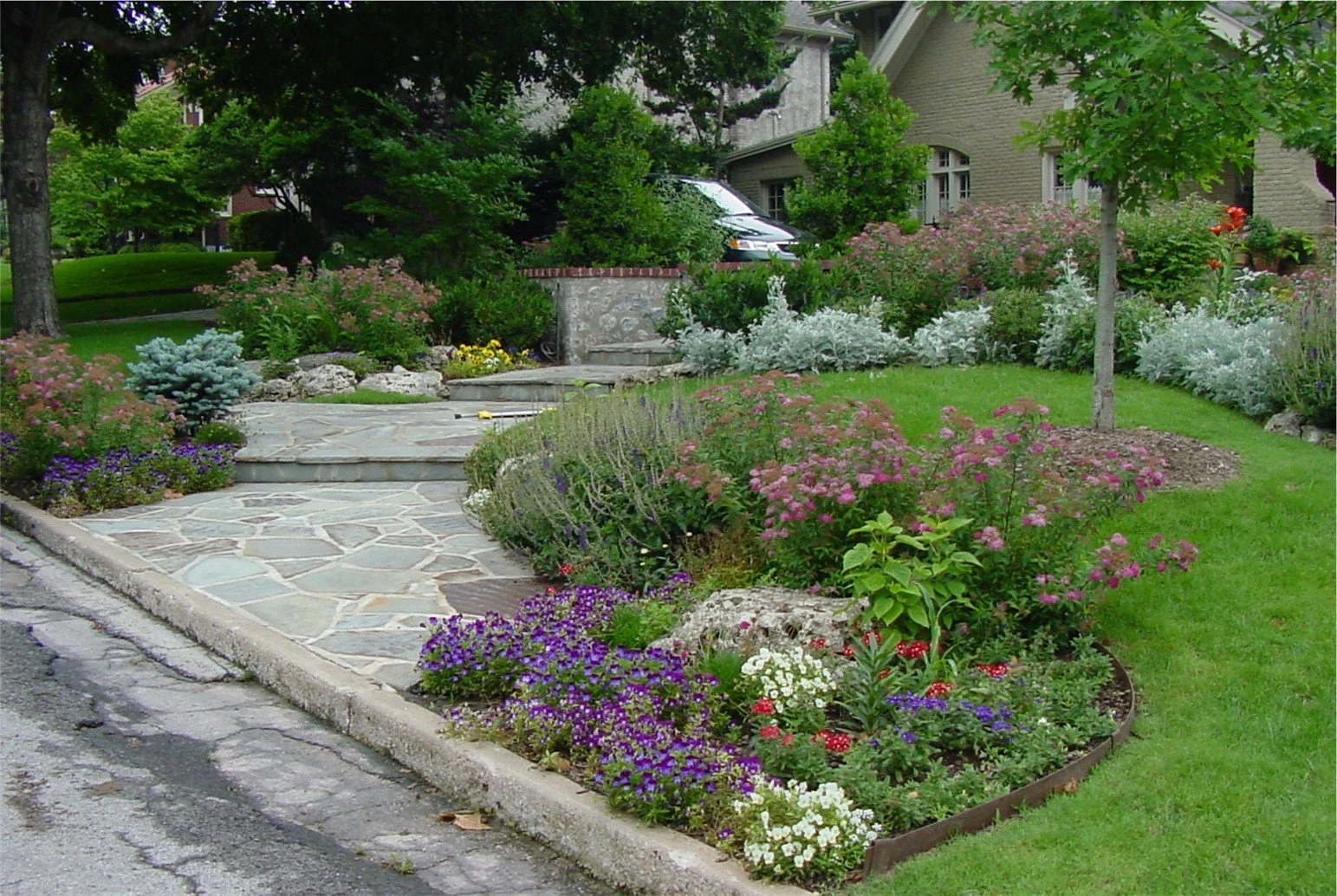 A lush front yard garden with colorful flowers, shrubs, and trees surrounding a stone walkway. A section of the street is visible in the foreground, with parked cars and residential houses in the background.