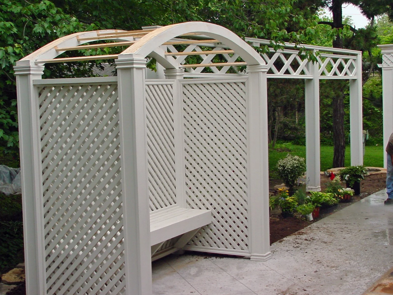 A white wooden garden arch with lattice panels and a bench, under construction, surrounded by green trees and plants.