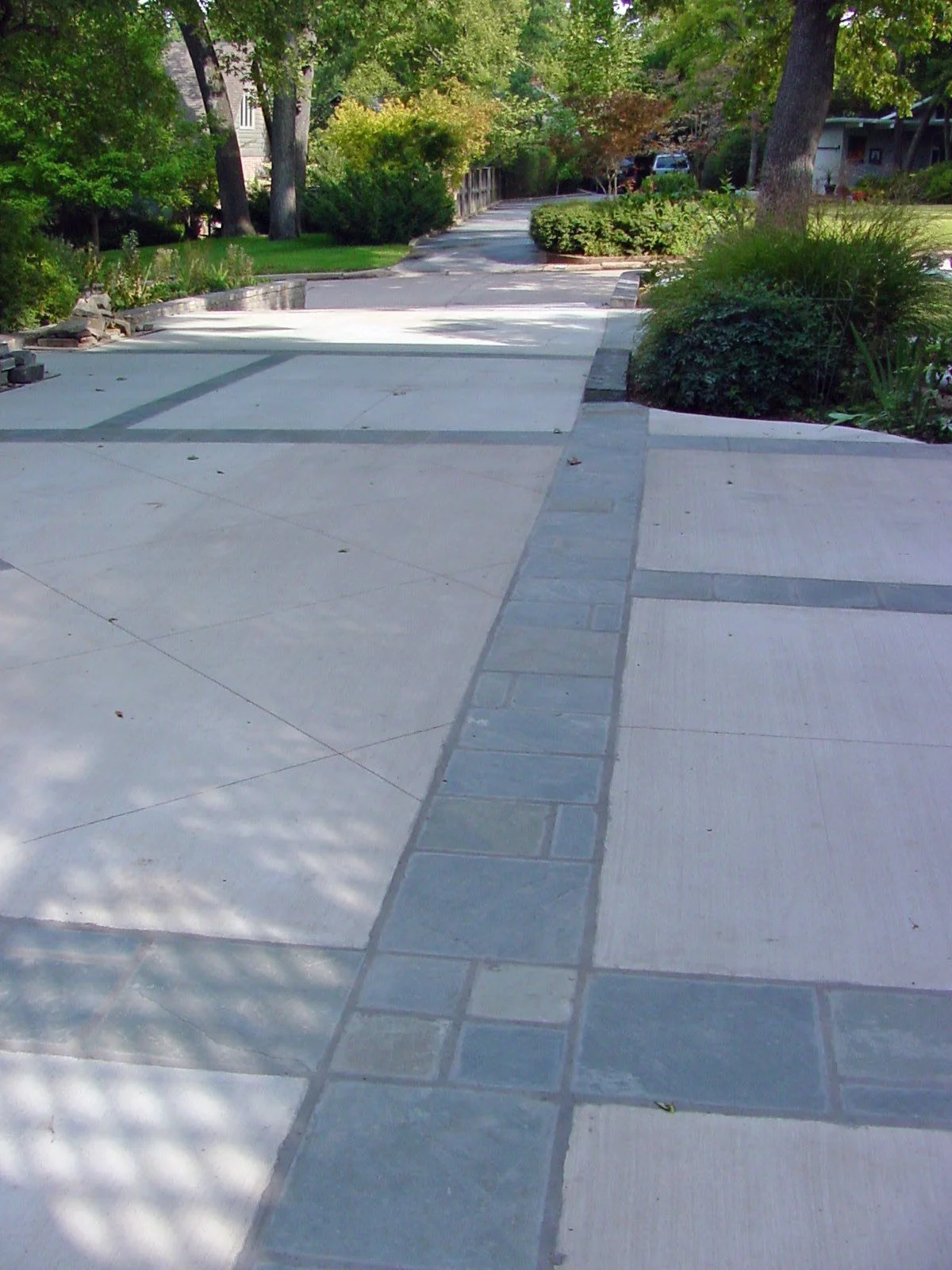 A paved sidewalk with a strip of darker tiles running down the center, surrounded by green bushes and trees, in a residential neighborhood.