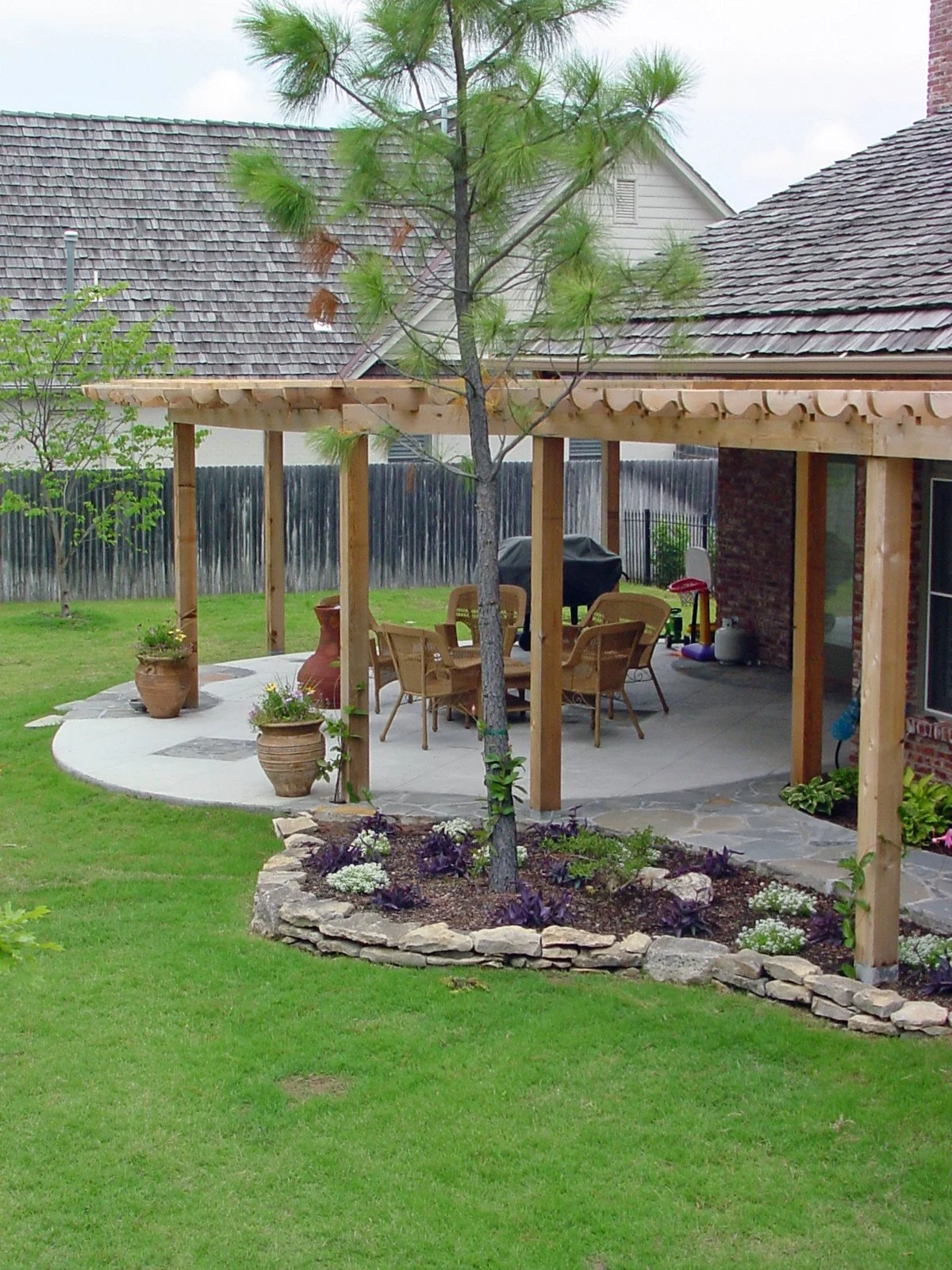 A backyard patio with a wooden pergola, surrounded by a grassy lawn and garden beds, with outdoor seating, potted plants, and a grill.