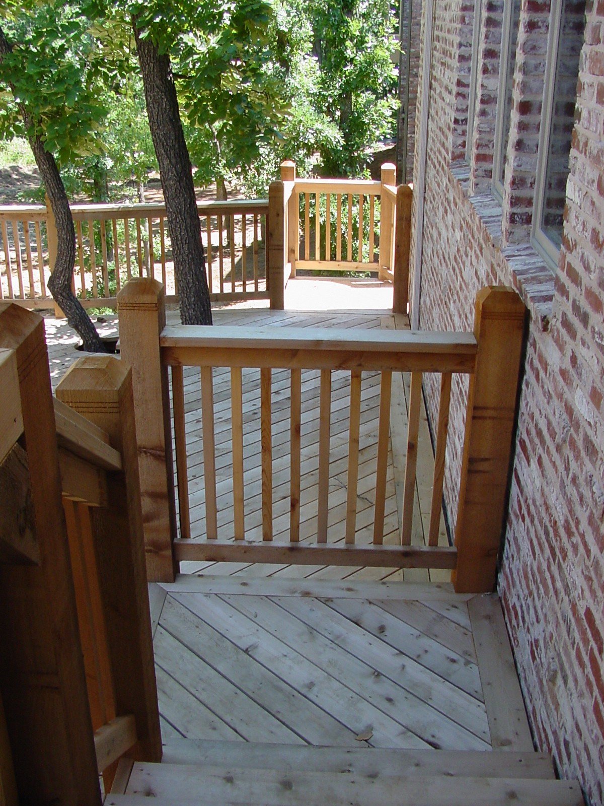 A view from a wooden porch with railing, adjacent to a brick house, overlooking trees in a backyard.