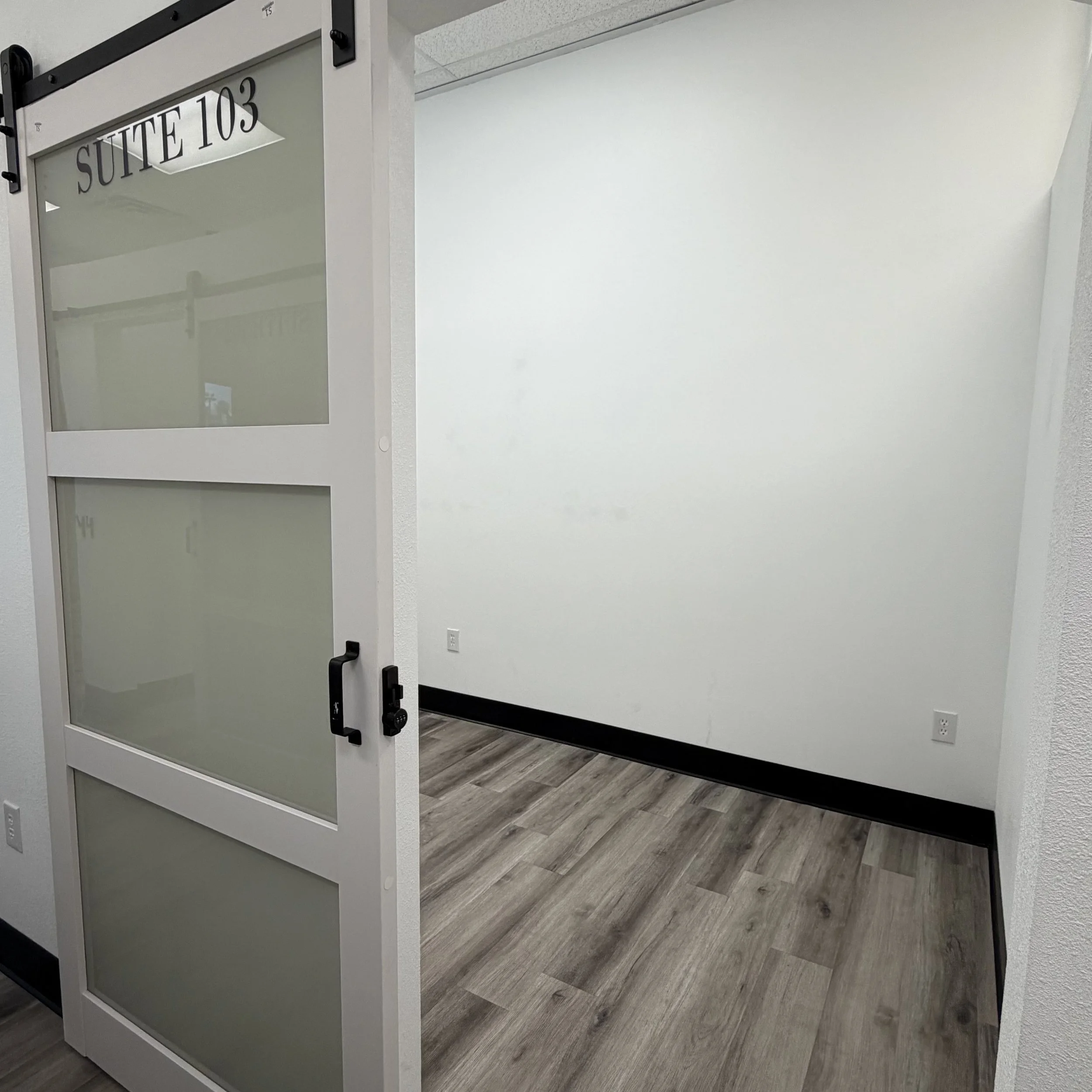 Office door with a glass panel labeled 'SUITE 103', open revealing a white wall and wooden flooring inside.