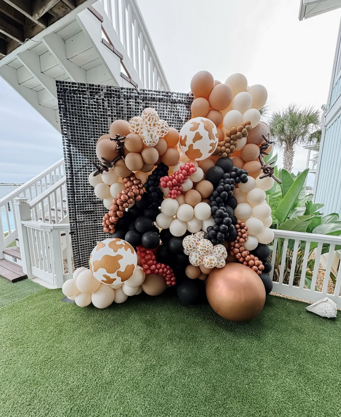 Decorative balloon display with black, white, tan, gold, and copper-colored balloons, some with cow-print patterns, arranged possibly for a celebration outdoors near a white deck and tropical plants.