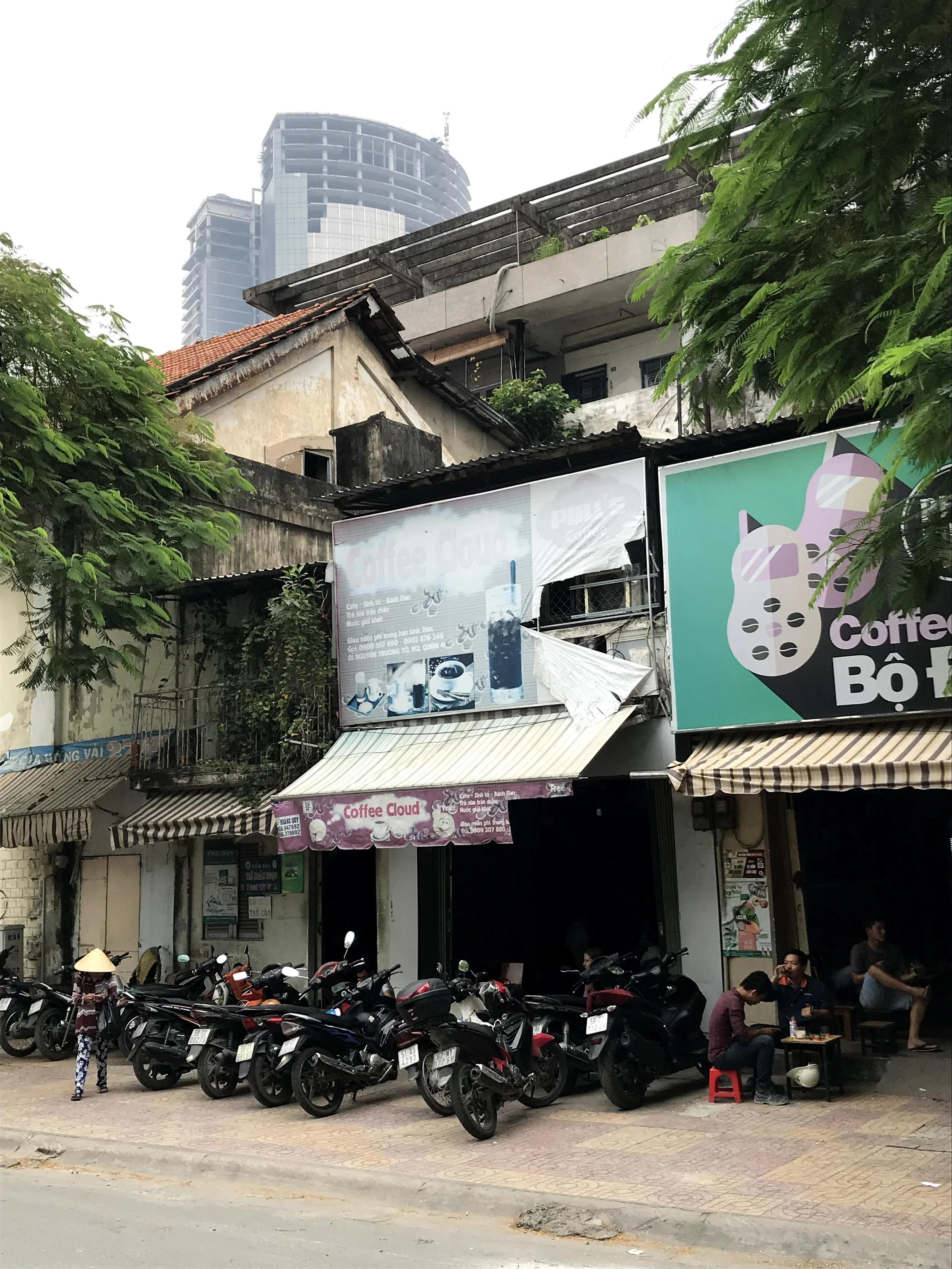 Street scene with parked motorcycles, a café with outdoor seating, people sitting and walking, trees, buildings in the background, and signs advertising coffee