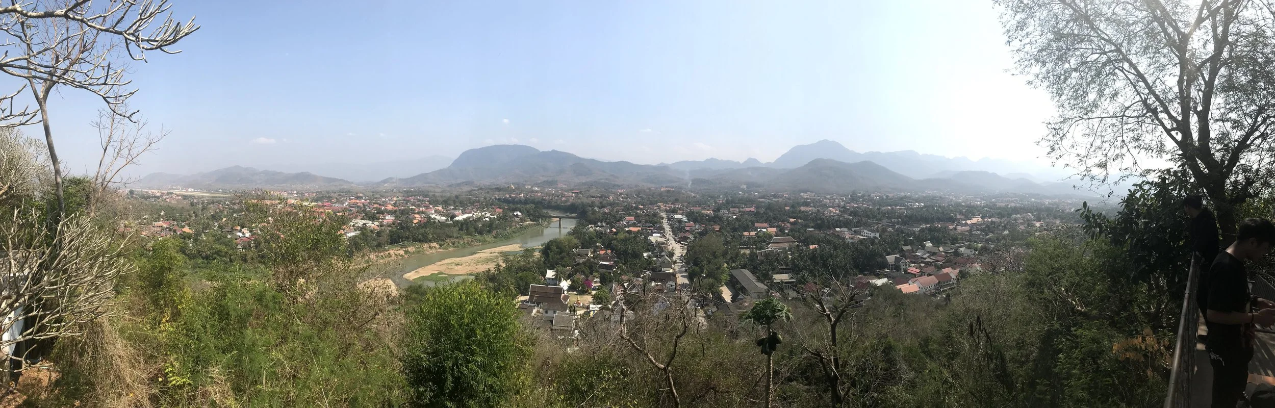 A panoramic view of a city overlooking a river, with houses, trees, and mountains in the distance under a clear blue sky.