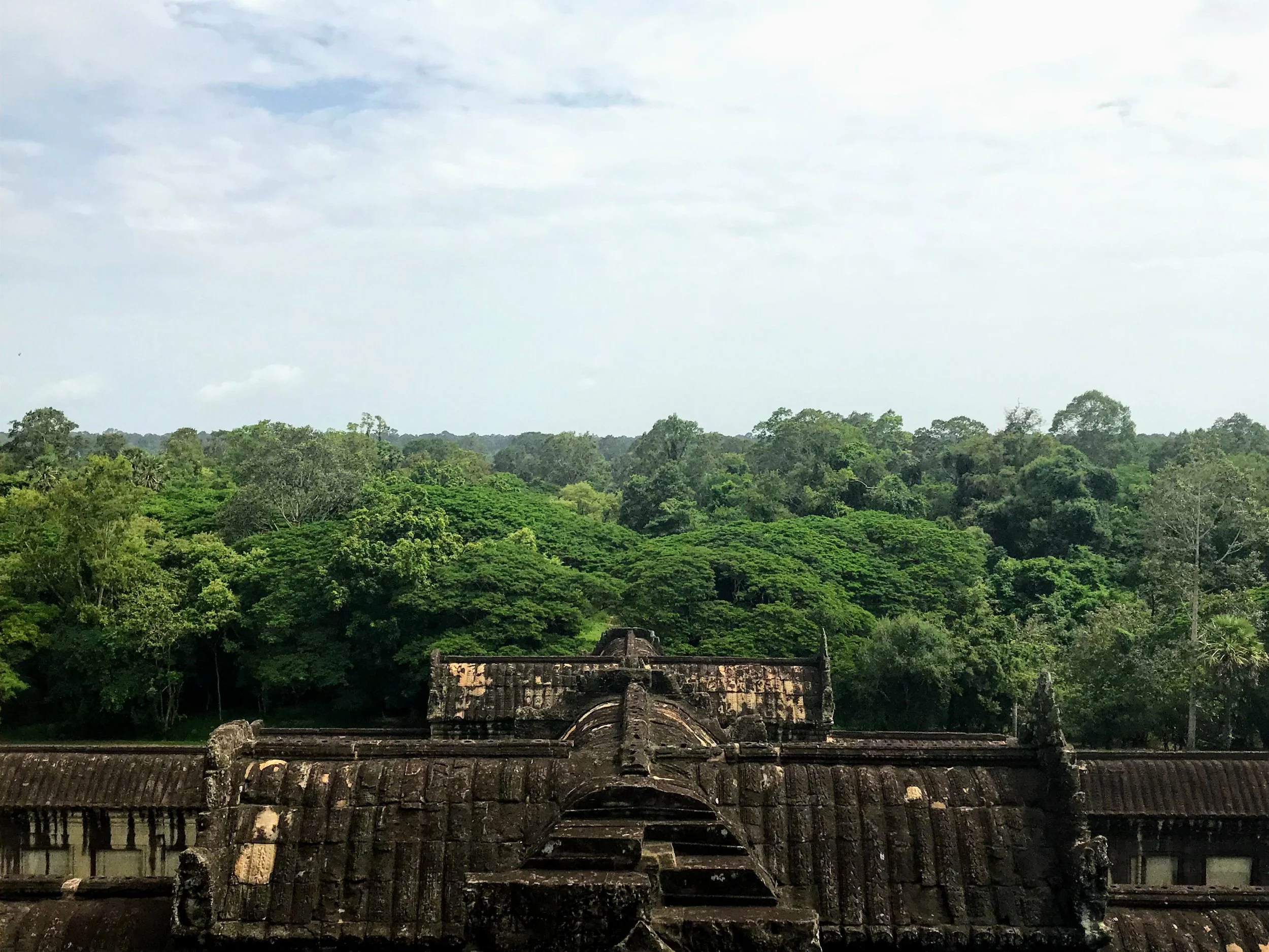 Ancient stone temple ruins at Angkor Wat, surrounded by a lush green jungle with a cloudy sky in the background.