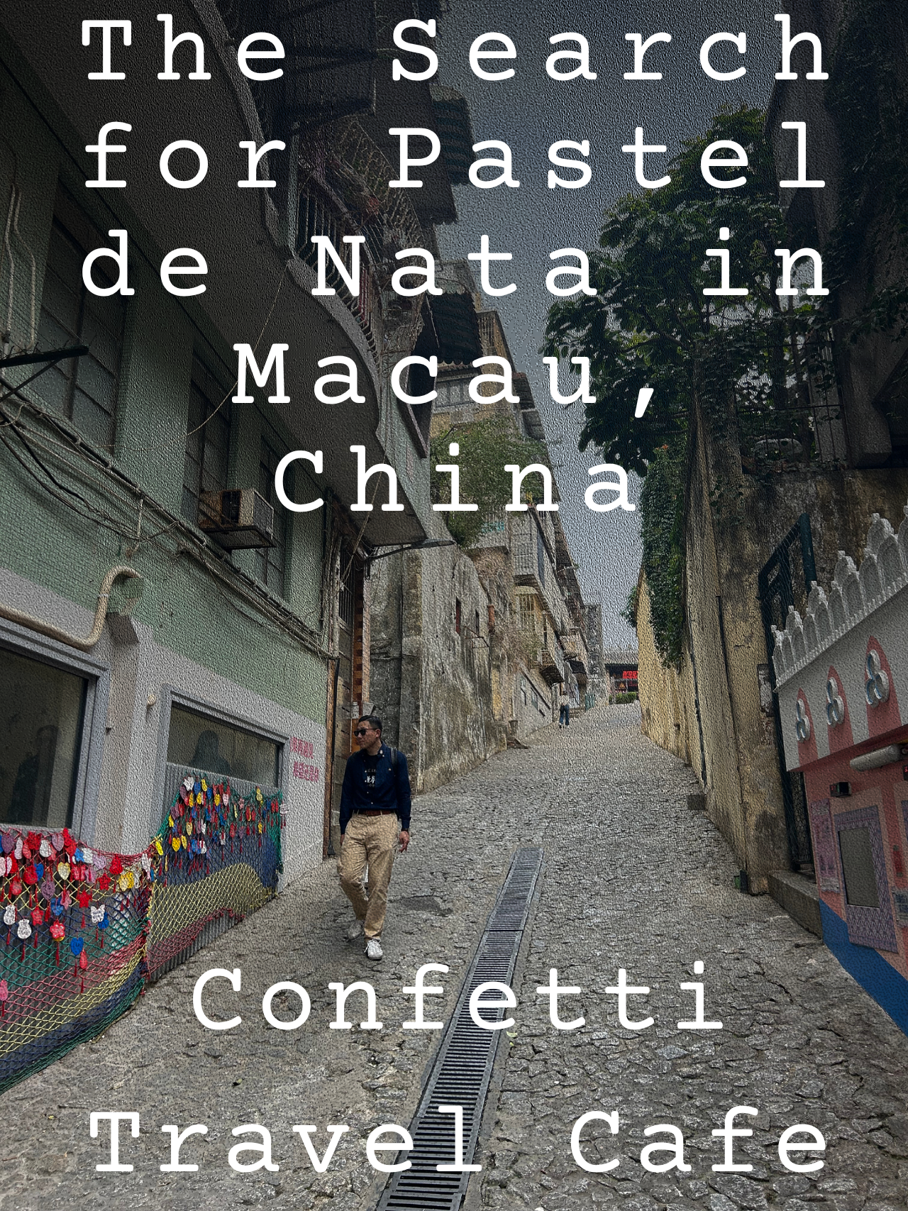 A narrow cobblestone street in Macau with buildings on either side. One man in sunglasses and casual clothing walks down the street, which curves uphill. There are colorful decorations on a fence on the left and trees with green leaves. Overlaying th