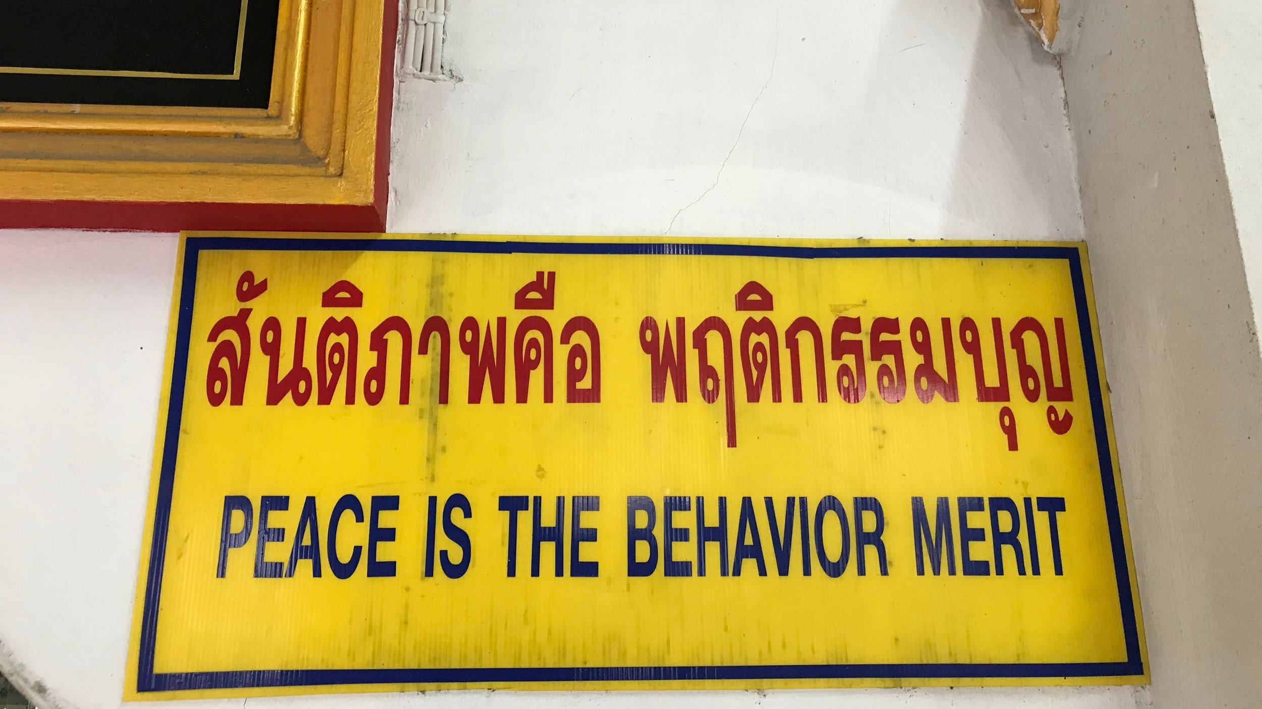 Yellow sign with red and blue text on a white wall, saying 'PEACE IS THE BEHAVIOR MERIT' in English and the same message in another language.
