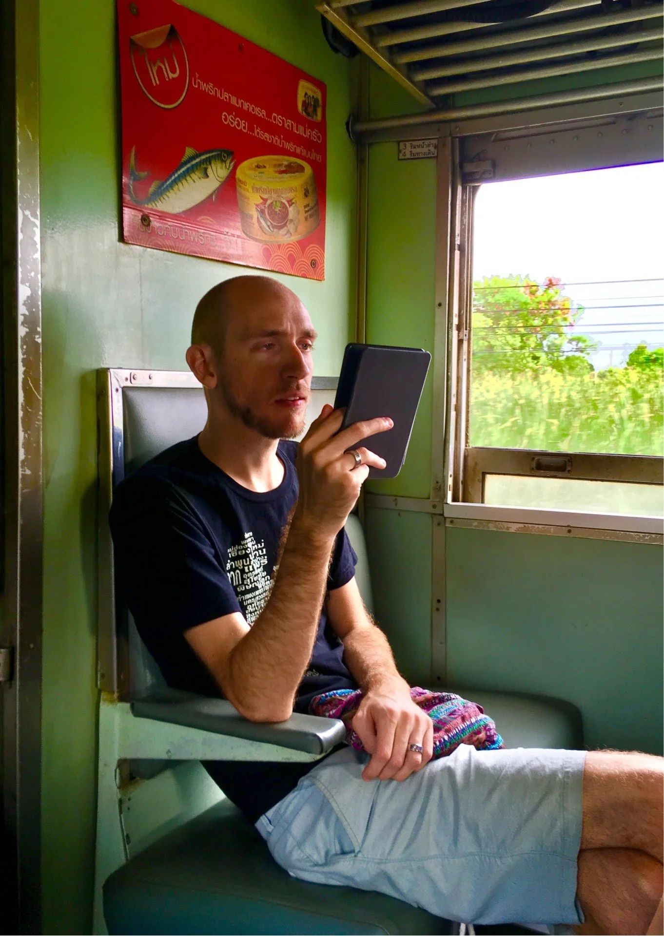 A man with light skin, a shaved head, and casual clothing is sitting on a train, holding a tablet, with a window showing trees outside. A colorful blanket is on his lap. There is a red advertisement with a fish and a can on the green wall behind him.