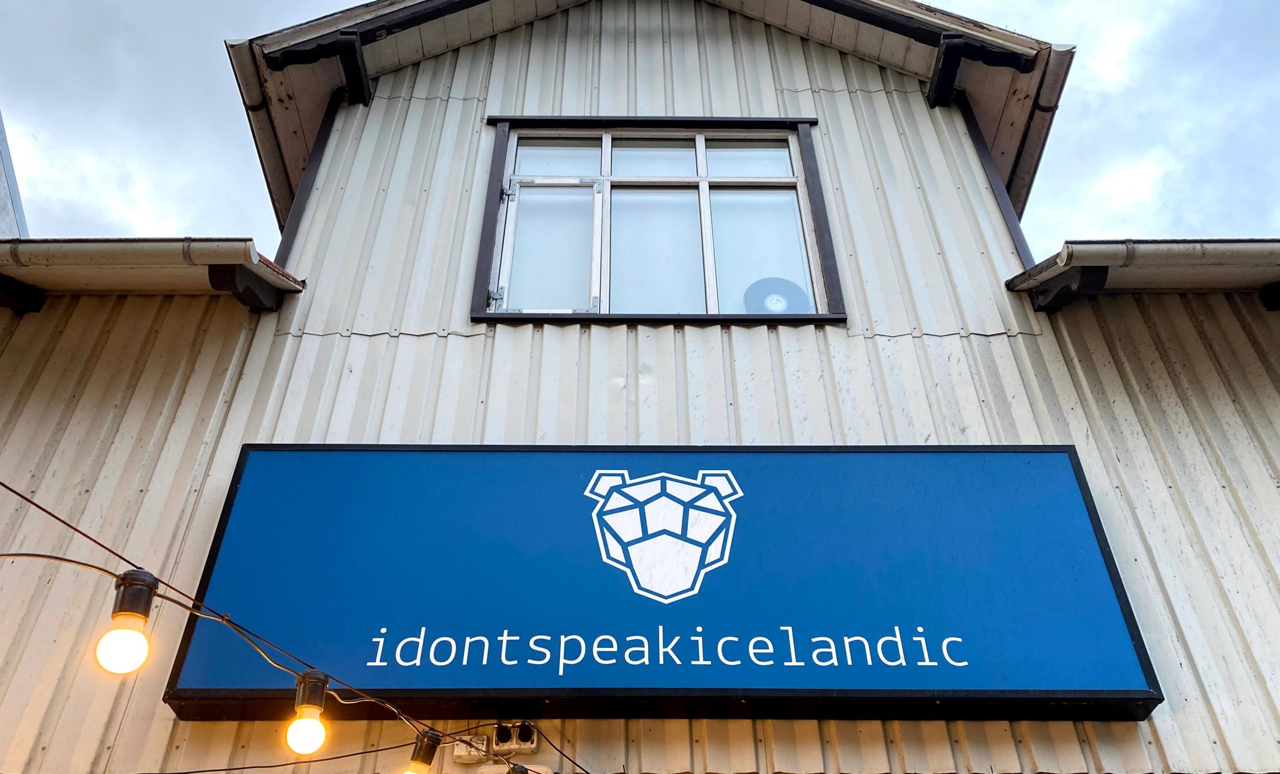 A building with a sign that reads 'idont speak icelandic' and a logo of a lion's head. The building has beige siding and a window with a metal frame. There are string lights with round bulbs hanging in front of the building.