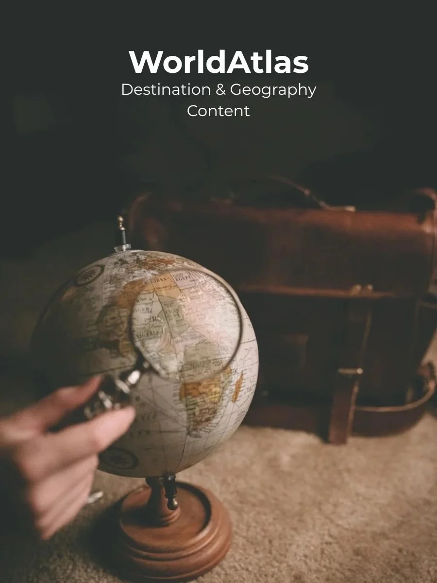 A person holding a magnifying glass over a globe, with a vintage trunk in the background and a dark wall, symbolizing exploration and geography.