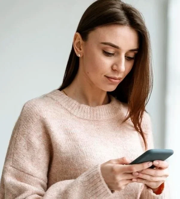 Woman with long brown hair looking down at her phone in a beige sweater indoors.