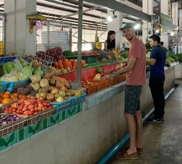 A man shopping for fresh vegetables at an outdoor market stand with a woman. The stand features an array of colorful produce including tomatoes, potatoes, peppers, apples, and onions.