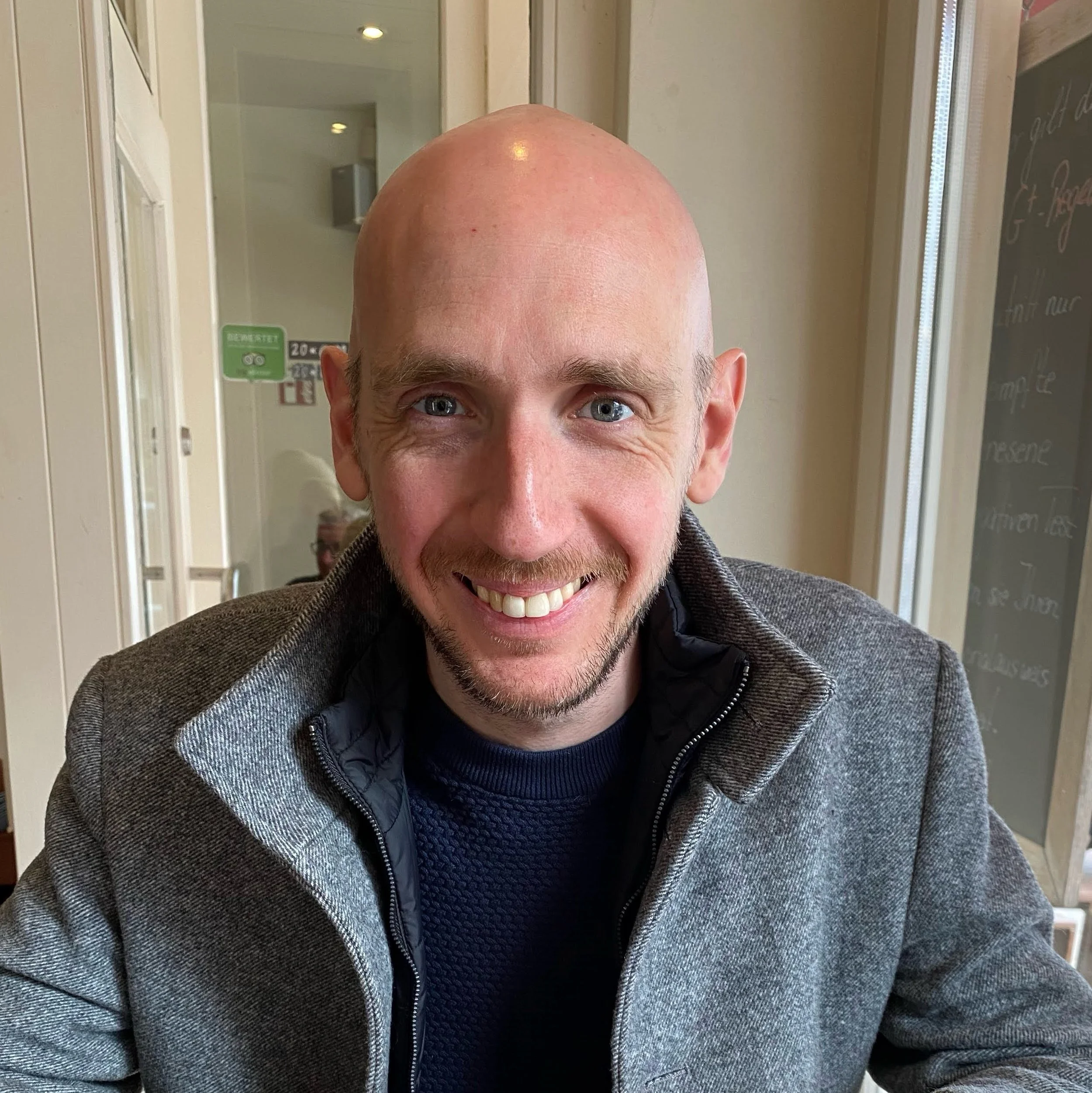 A man with a bald head, blue eyes, and a light beard smiling, wearing a gray textured jacket over a black shirt, indoors near a chalkboard and glass door.