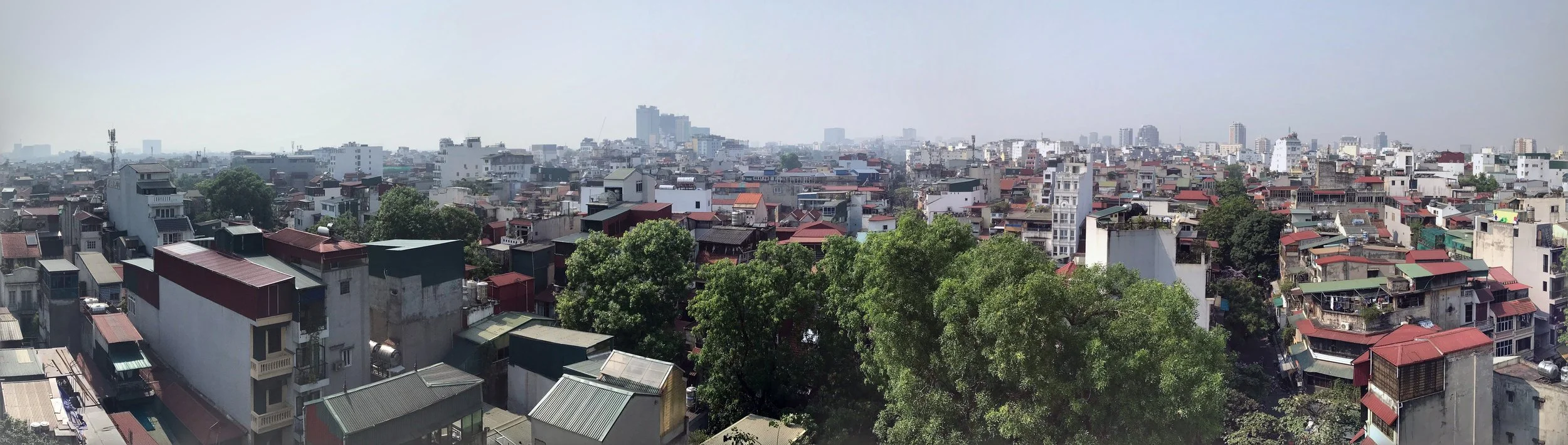 Panoramic view of a dense urban area with numerous buildings, top trees, and a hazy sky.