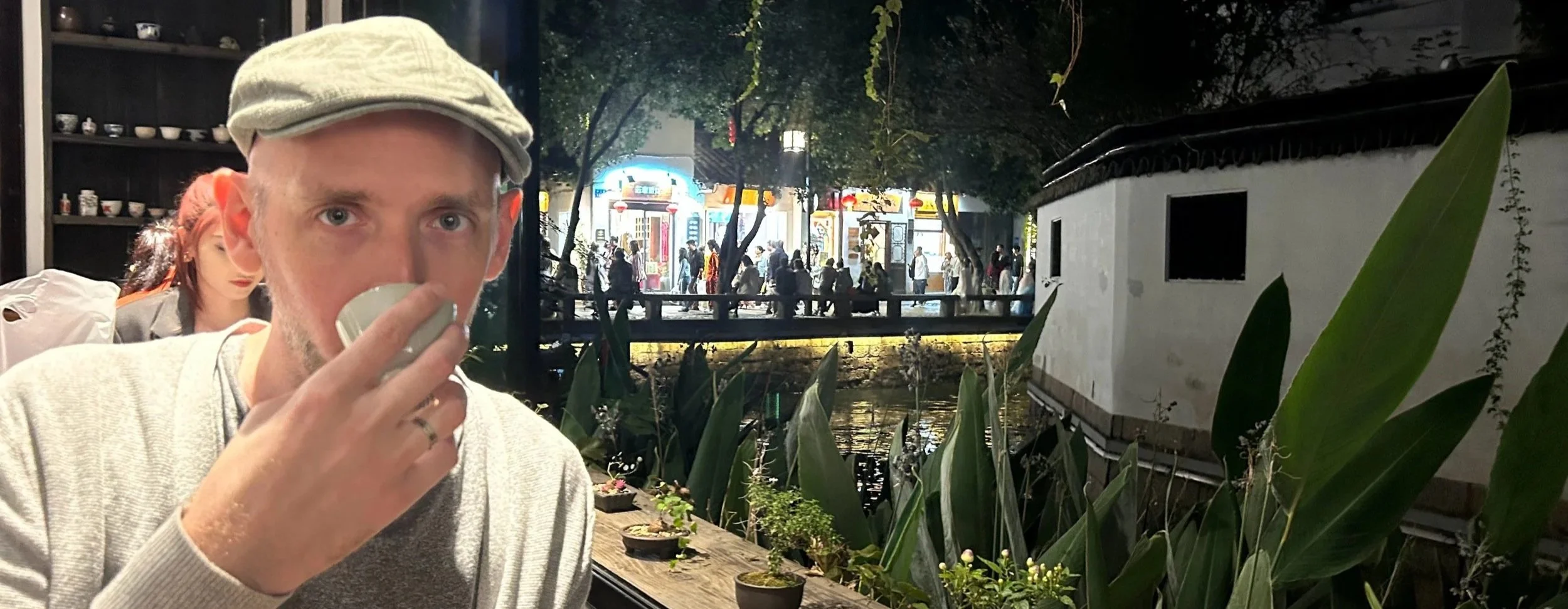A man drinking from a cup at a cafe with a night scene outside showing a street with people, lights, and trees.