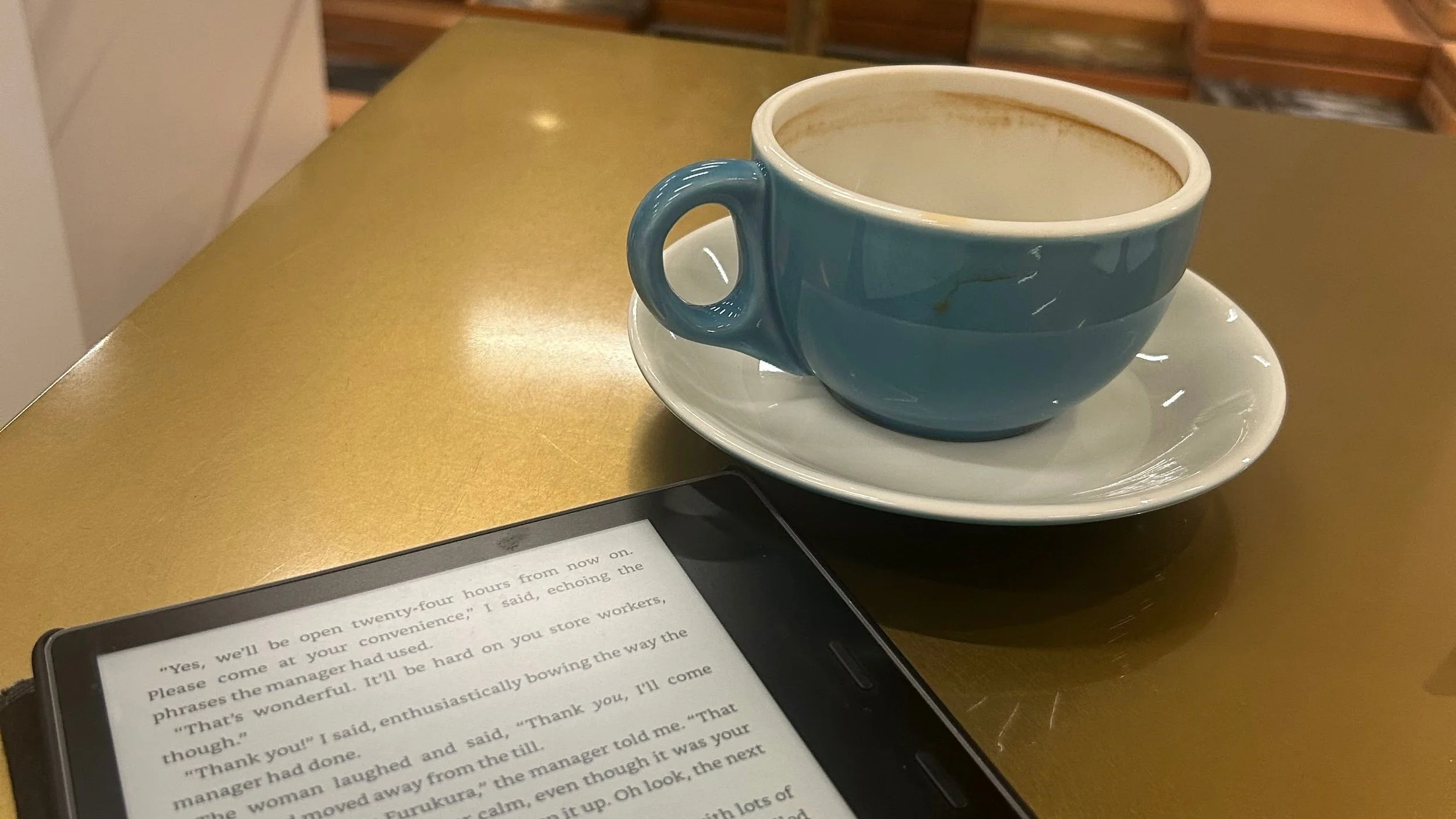 A blue ceramic cup with coffee or tea on a white saucer sitting on a gold-colored tabletop. A digital device displaying text of a story is partially visible at the bottom of the image.