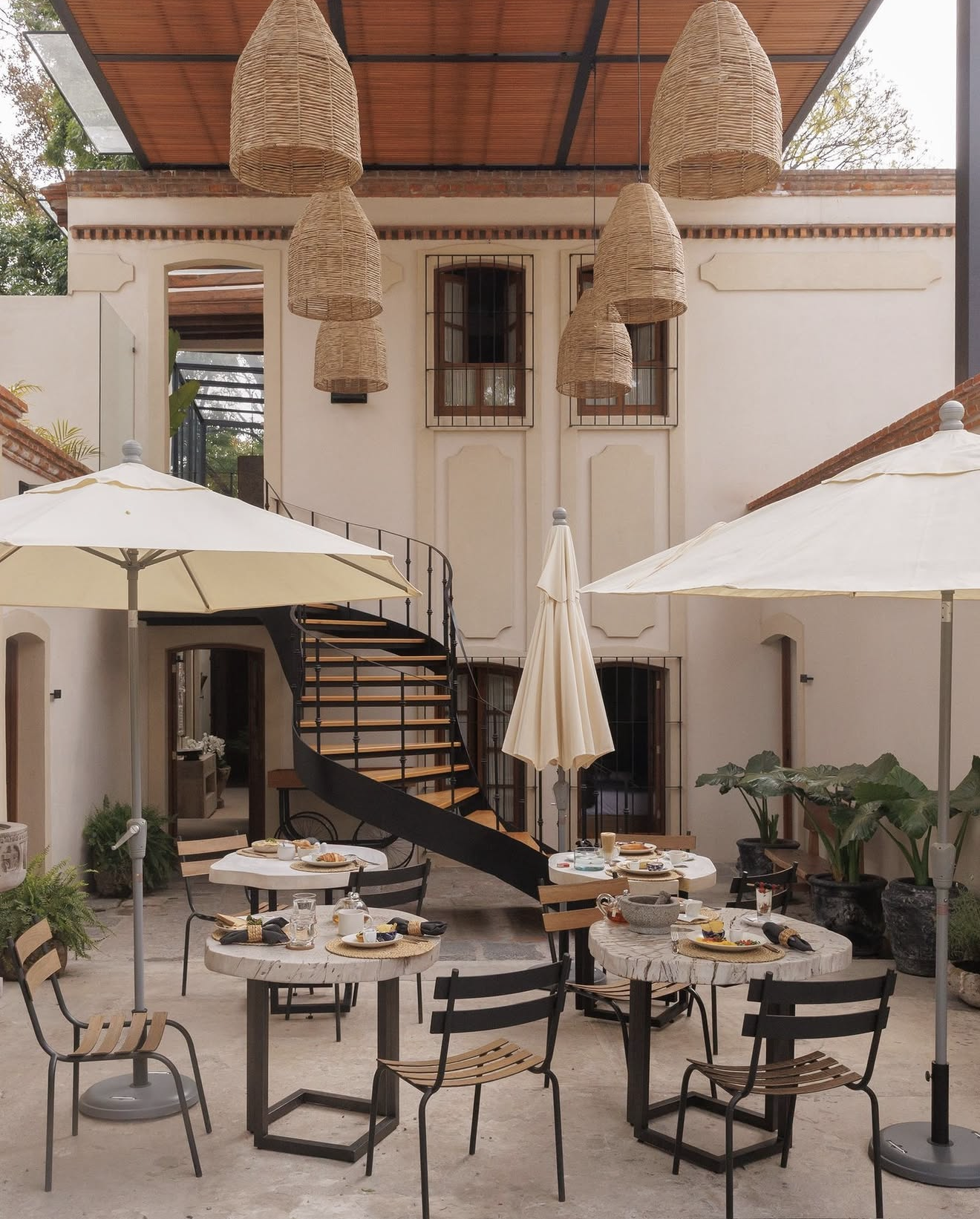 Outdoor patio with tables set for dining, large white umbrellas providing shade, a spiral staircase, and hanging wicker light fixtures