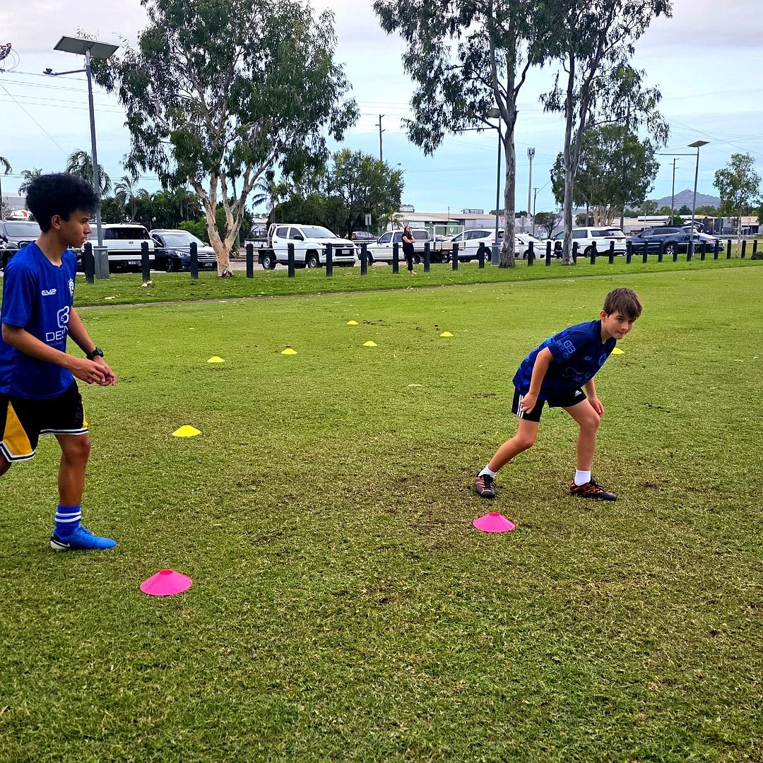 Two boys training on a grassy field with yellow and pink cones, while a woman watches in the background.
