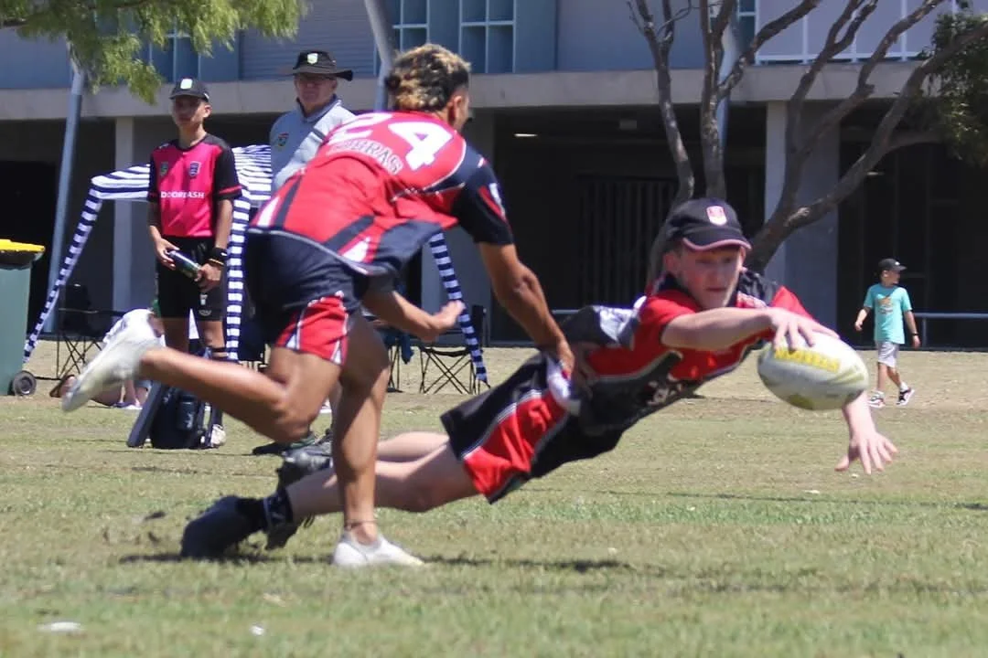 A young boy in a black and red rugby uniform is diving forward to catch a rugby ball during a game, with a teammate in a matching uniform chasing him from behind on a grassy field.