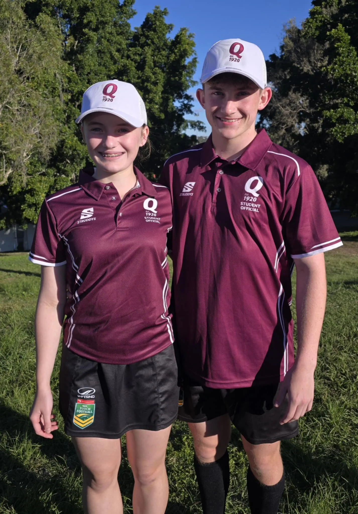 Two teenagers wearing maroon and white sports uniforms and white caps standing outdoors on grass, smiling at the camera.