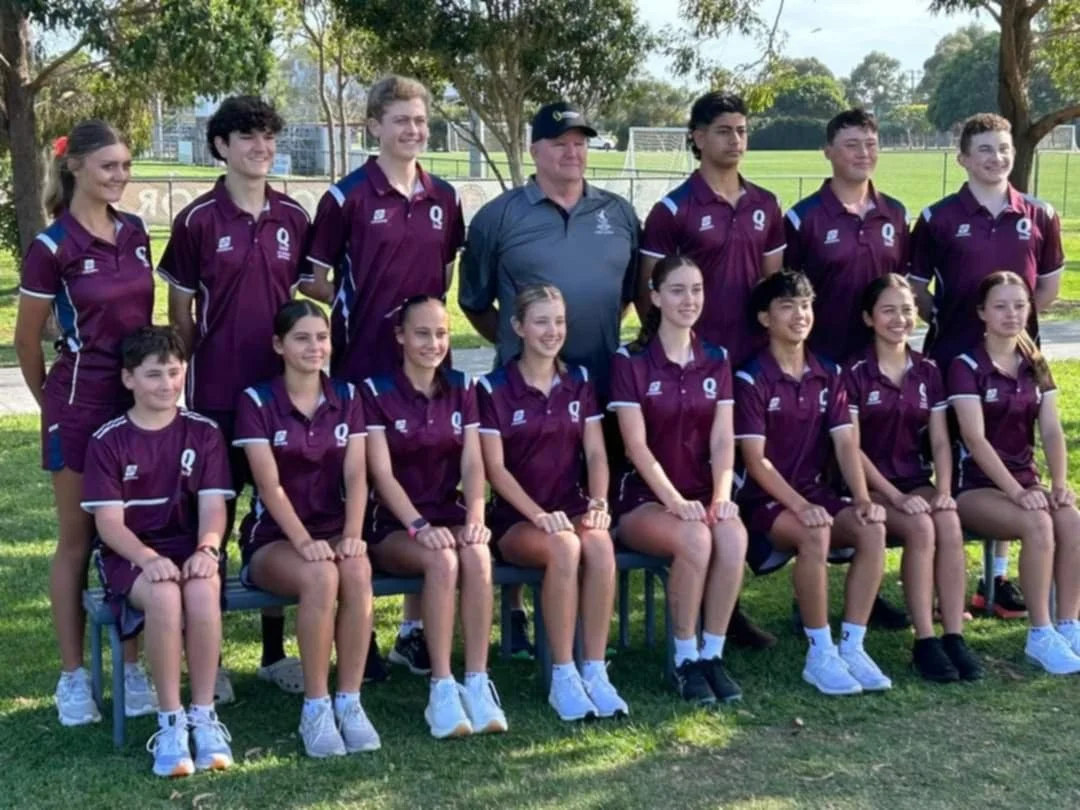 A group of young girls and boys in sports uniforms posing outdoors with a coach. The team is dressed in matching maroon jerseys with blue and white accents, sitting and standing for a team photo on a grassy field with trees in the background.