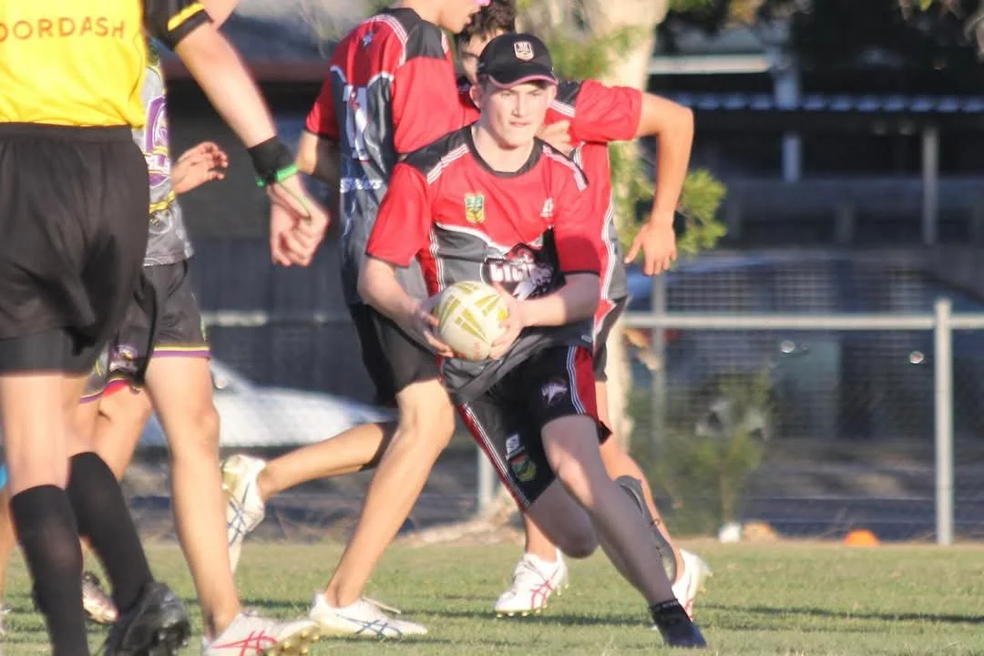 A young rugby player wearing a red and black uniform holding a rugby ball while running on the field during a game.