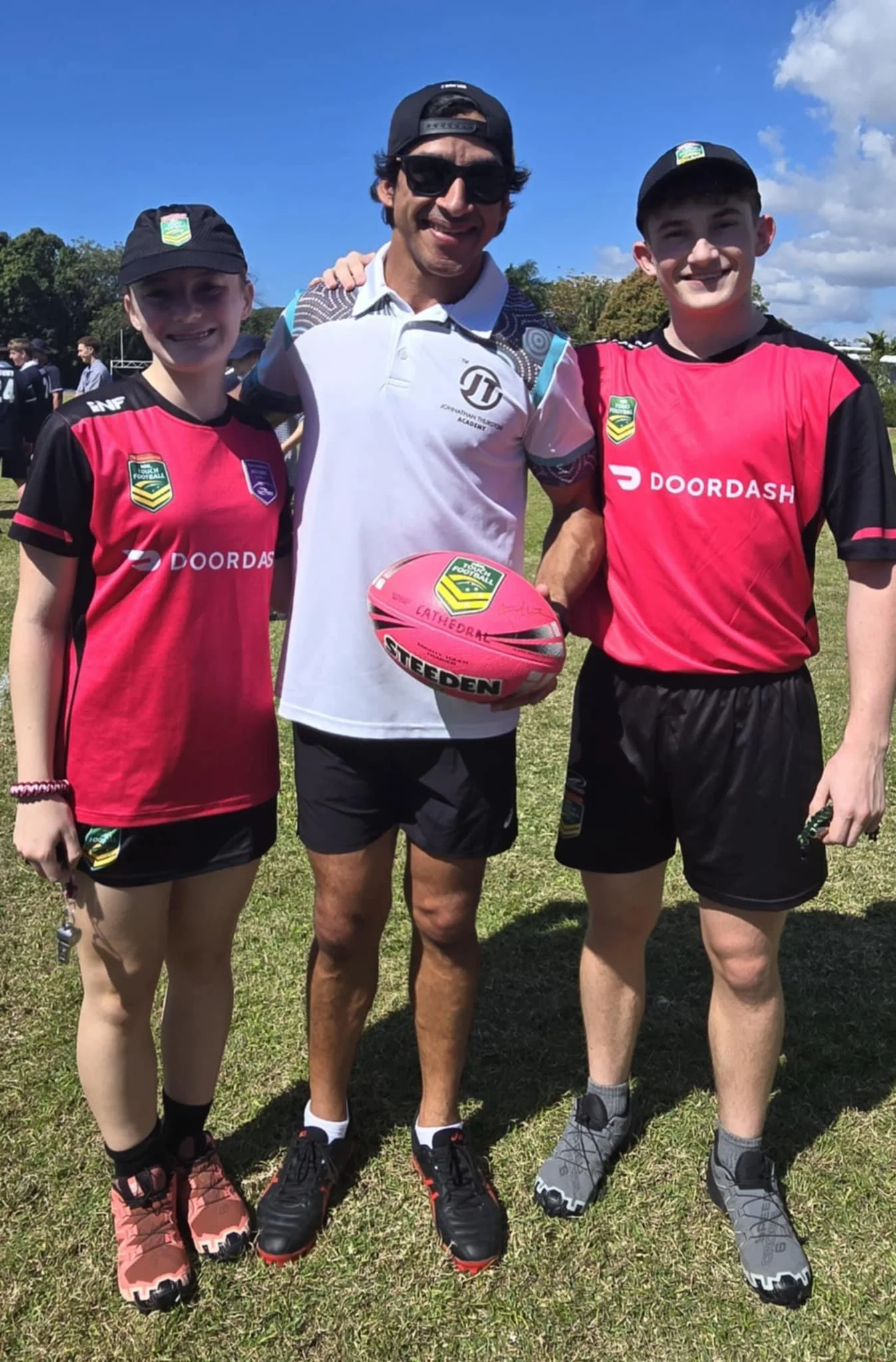 Three people standing on a grass field, two young athletes in pink and black sports uniforms and an adult man holding a pink football. The sky is blue with some clouds.