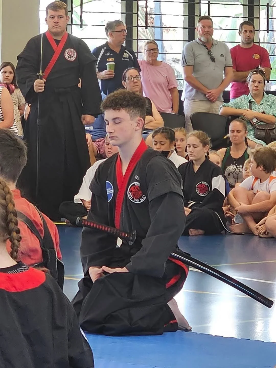 A young martial artist in a black uniform with red accents, sitting in a martial arts pose, in a martial arts class or competition setting. Several children and adults are seated or standing around, some watching attentively, in a room with large windows and a blue mat on the floor.