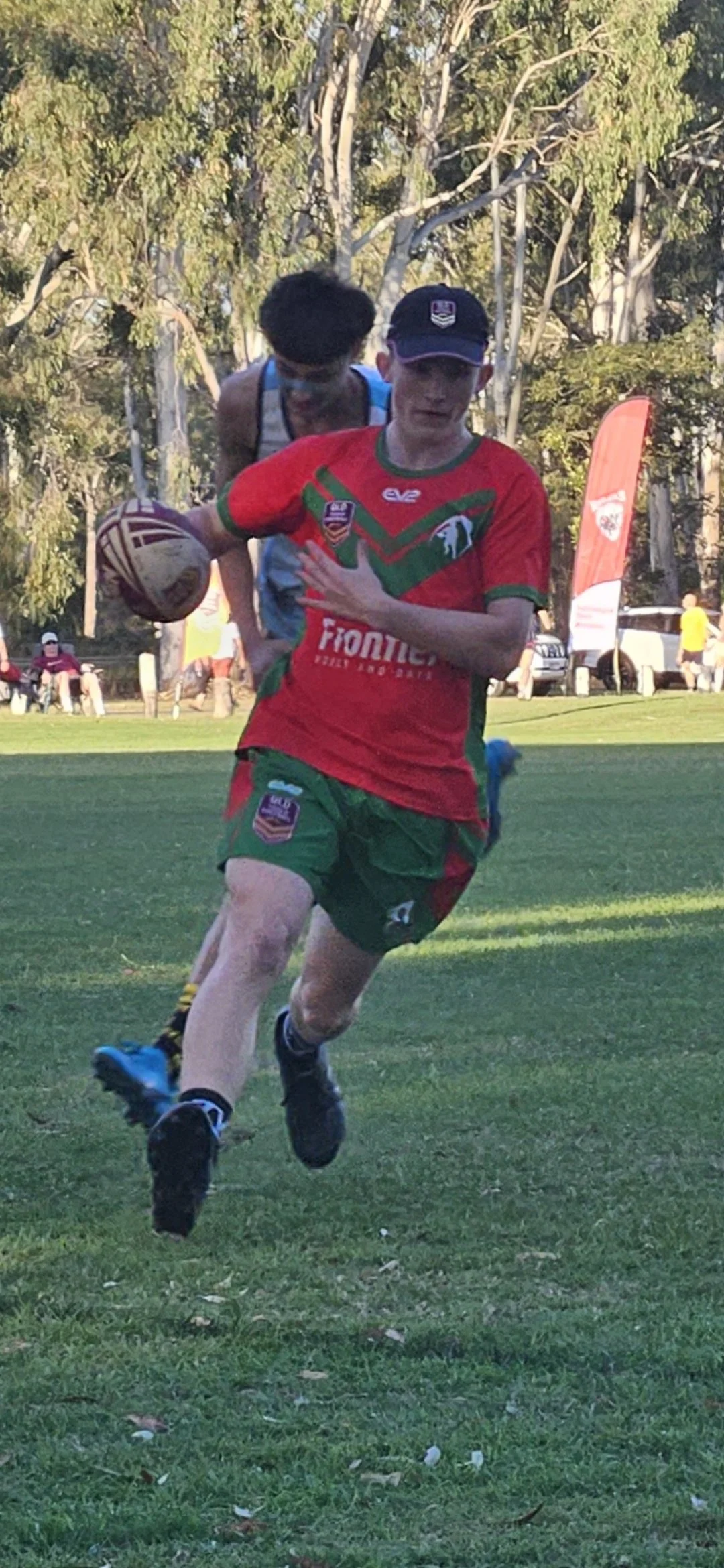 A young boy in a red and green sports uniform running with a rugby ball on a grassy field, with trees and spectators in the background.