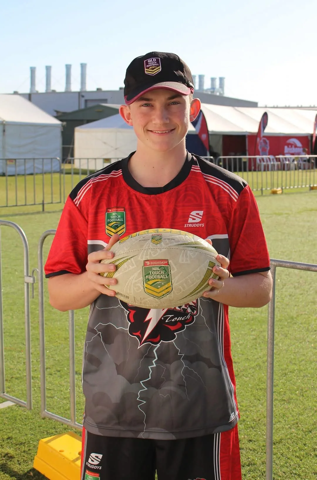 Young male athlete in a red and black sports jersey holding an rugby ball, standing outdoors on a grass field with event tents and barriers in the background.