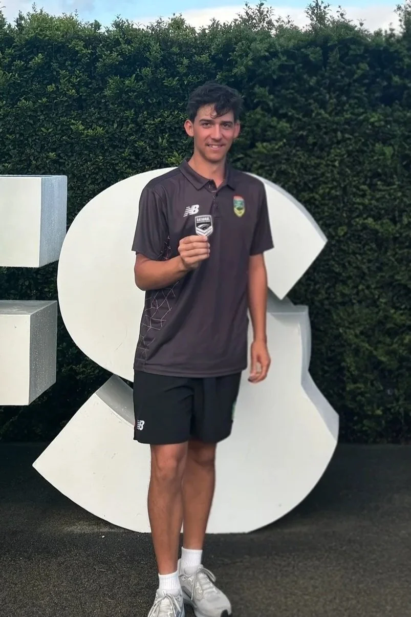 A young man in black athletic clothing holding a sports team badge or patch, standing outdoors in front of large white decorative letters or symbols and greenery.