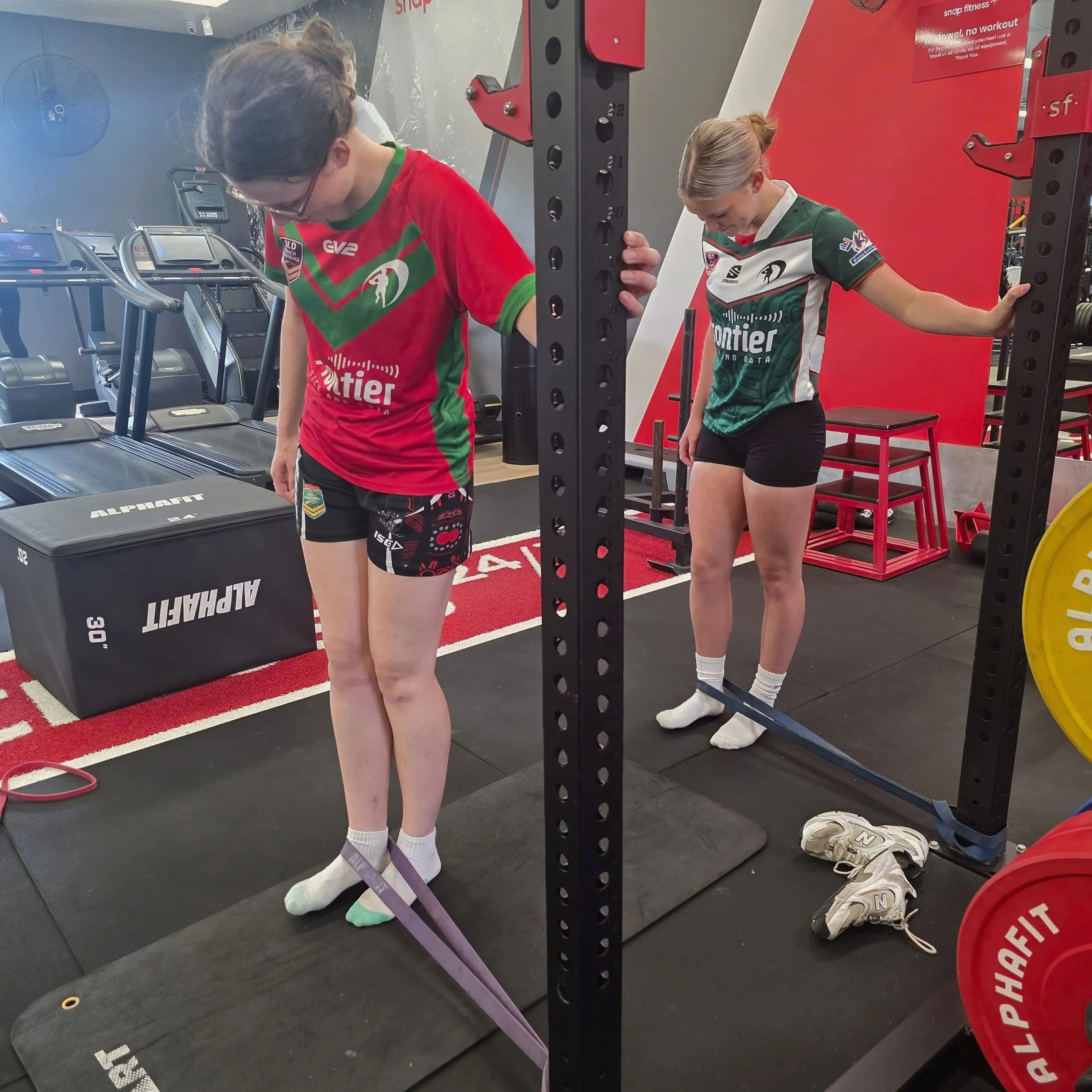 Two women in athletic clothing are standing on treadmill machines in a gym. One is wearing a red and green shirt, black shorts, and glasses, looking downward. The other has blonde hair tied back, wearing a green, white, and black shirt, and is looking down with her right arm resting on a piece of gym equipment. There are weight plates, shoes, and other gym equipment around them.