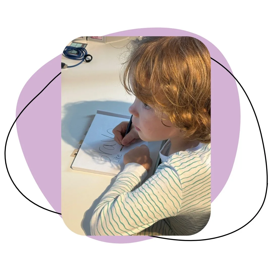 A young boy with curly red hair is sitting at a white desk, writing in a notebook with a black marker. The notebook has the word 'BRAVE' written on it, and the boy is in a bright space with some electronic cords on the desk.