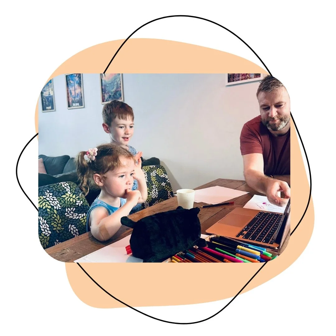 A man and two young children sitting at a wooden table, with art supplies and a laptop, in a room with framed pictures on the wall.