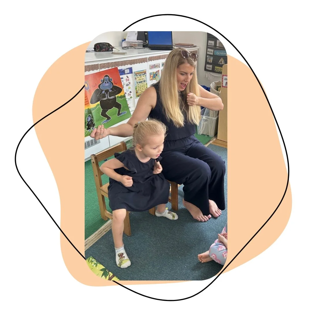 A woman and a young girl sit on small chairs in a classroom, engaging in a playful activity. The girl is leaning forward while the woman appears to be in mid-movement, both smiling and enjoying the moment. The classroom features colorful posters and educational materials on the walls.