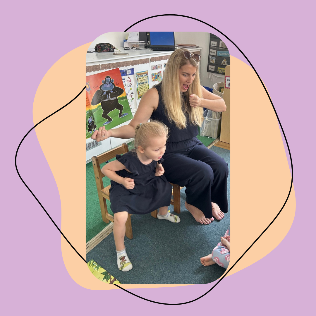 A woman and a young girl are sitting on chairs in a classroom, appearing to sing or act out a story together. The woman is holding a picture book, and both are dressed in black. The classroom has colorful educational posters and materials on the walls.