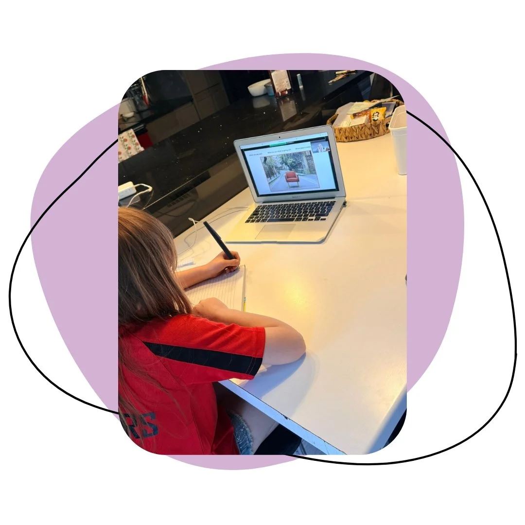 A child in a red shirt doing homework at a white table, with a laptop showing a picture of a chair and window, in a kitchen setting.