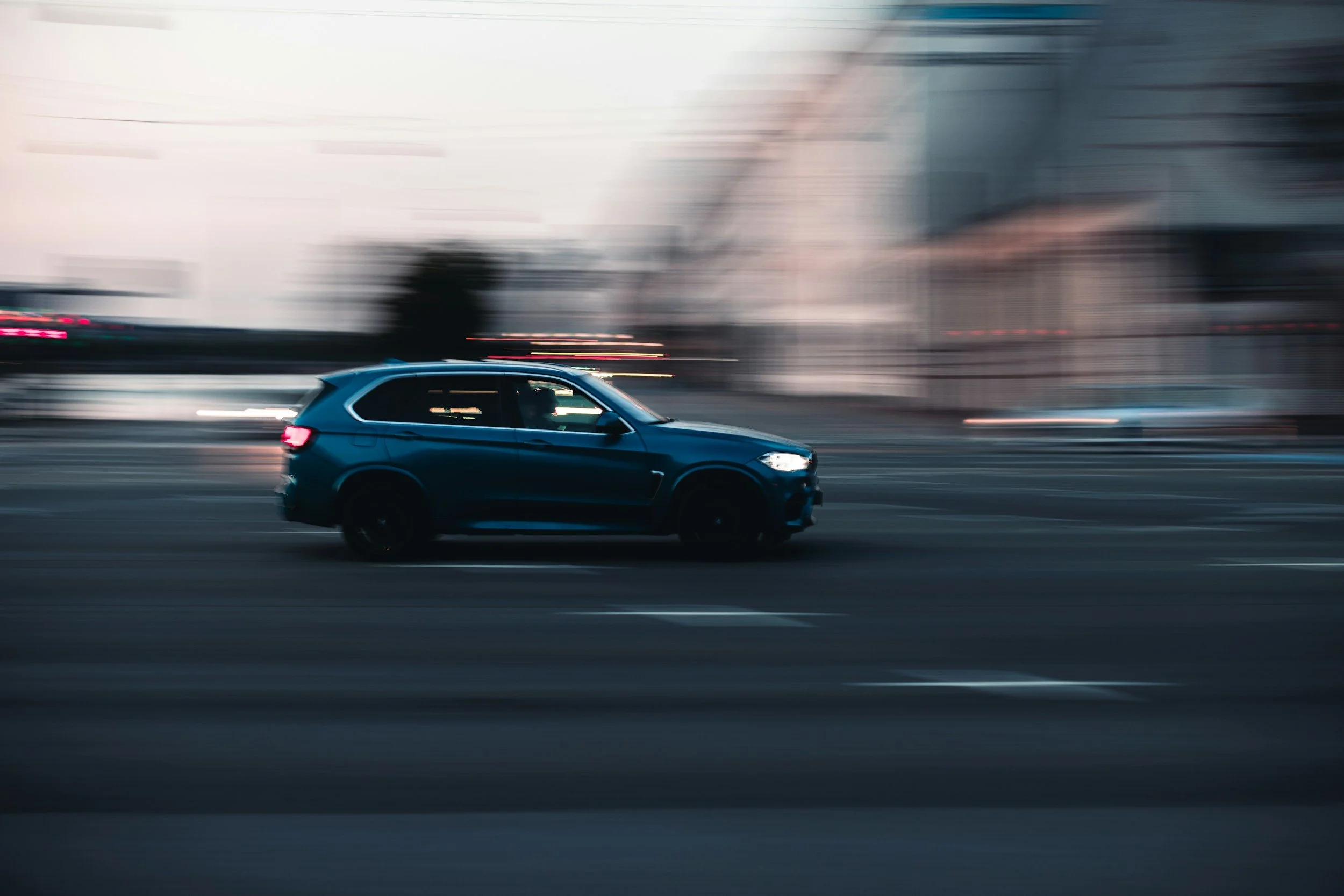 En blå SUV kører hurtigt gennem en bygade under skumring, med den bagvedværende bys blokke og lys der er slørede på grund af bevægelsen.