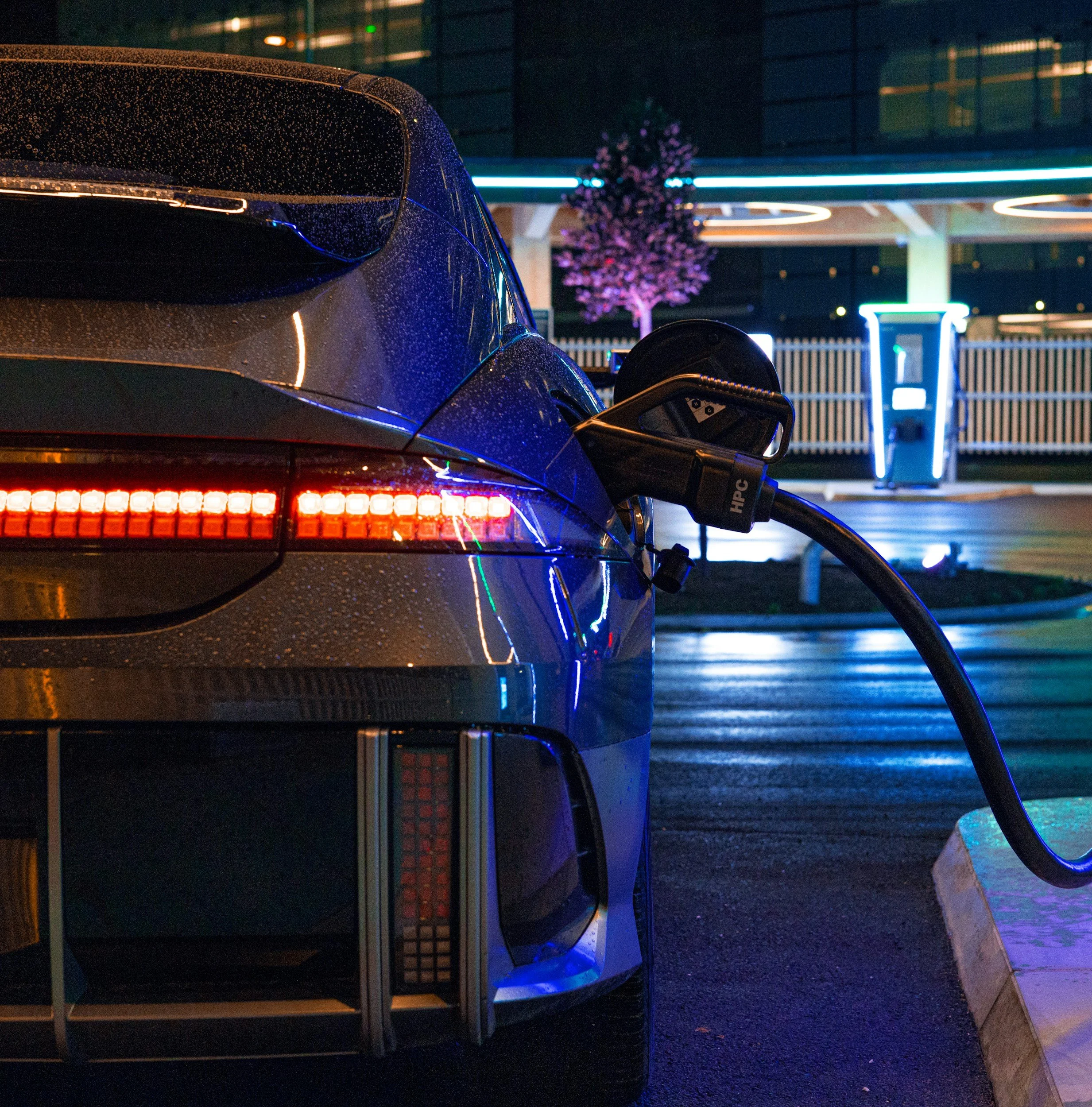 Natten, en elbil bliver opladt ved en ladestation med neonlys og en blomst i baggrunden