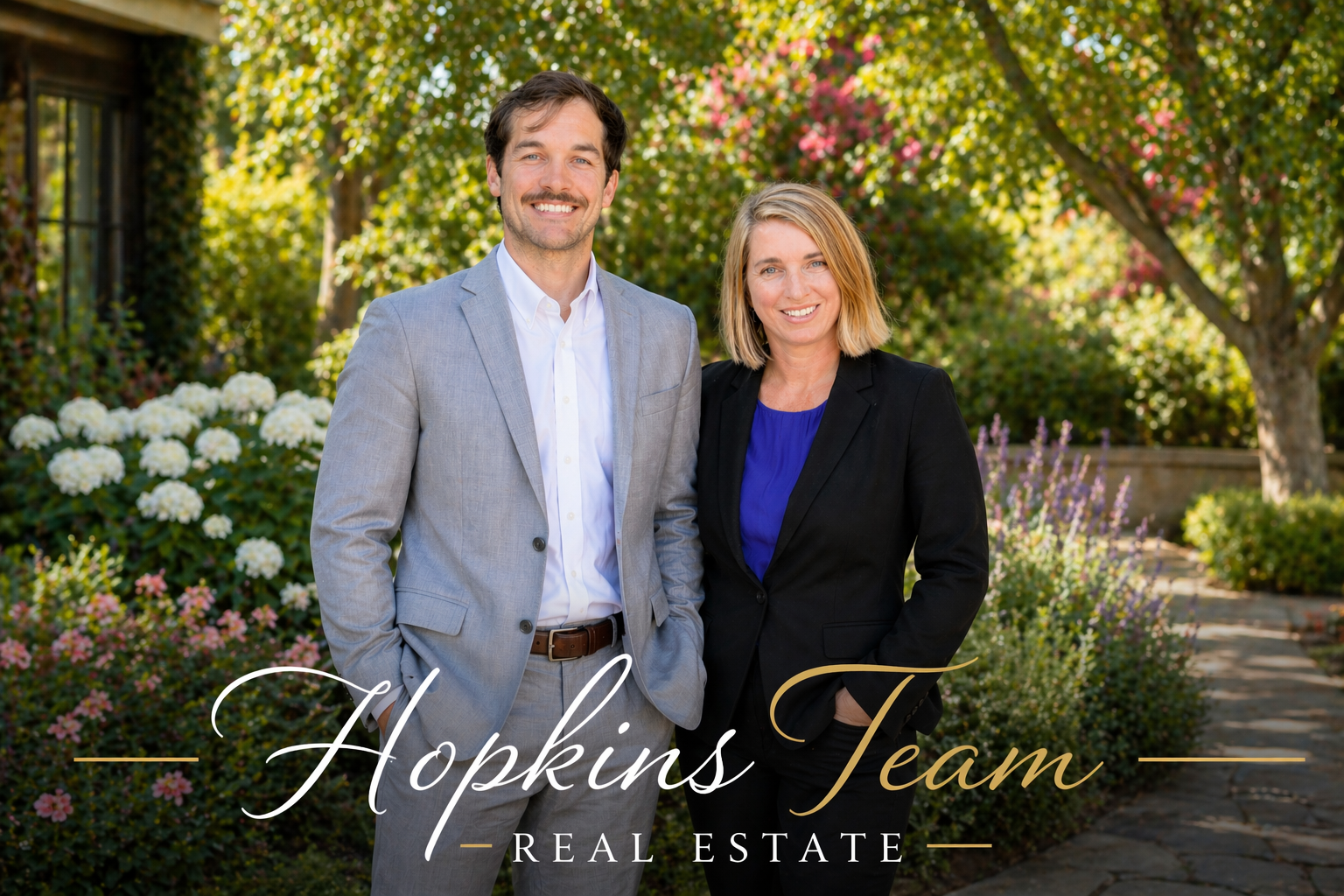 Hopkins Team Real Estate agents standing outdoors in a landscaped garden, featuring a professionally dressed man in a light gray suit and a woman in a black blazer, smiling in a bright residential setting.